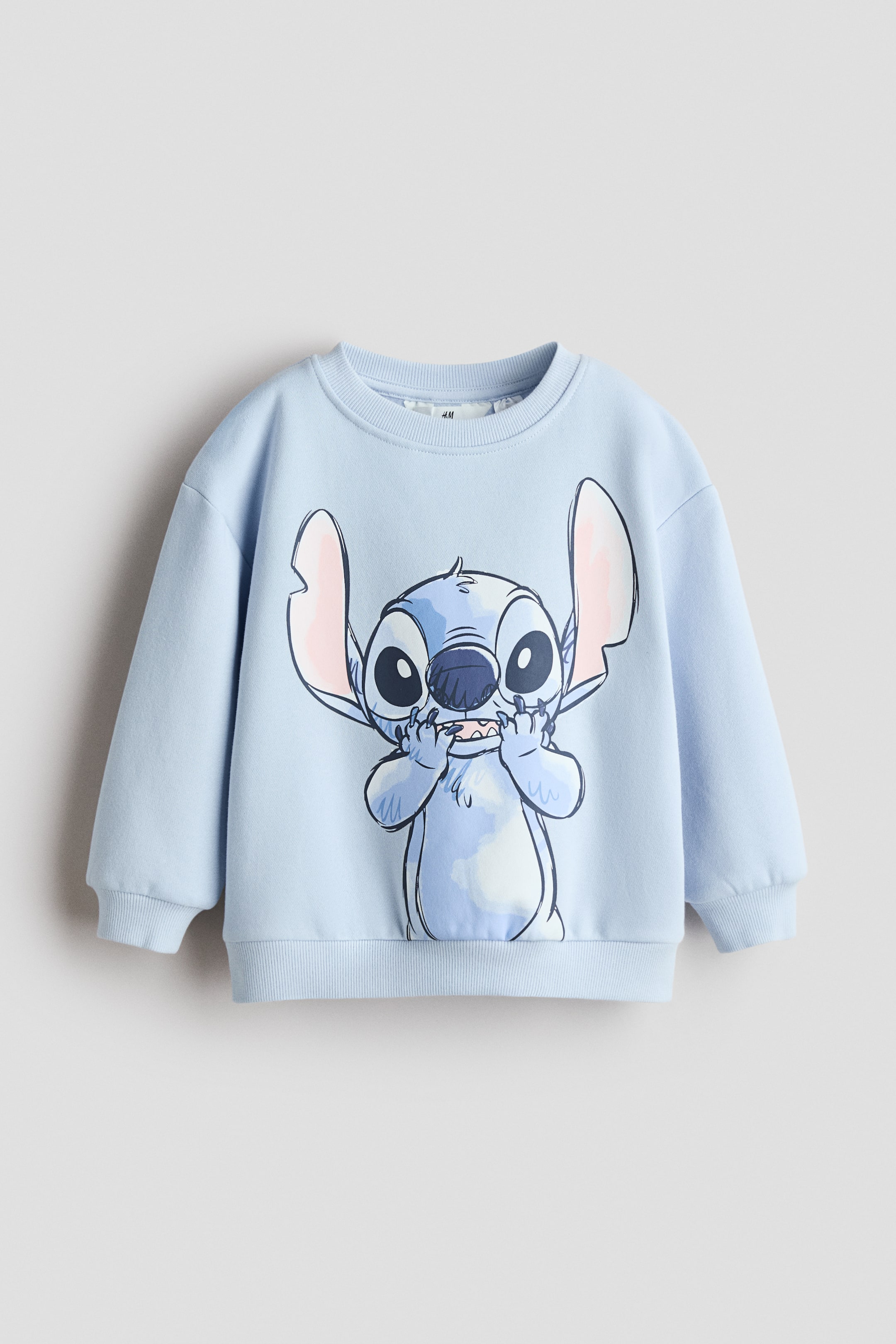 View larger image: This light blue crew neck sweatshirt showcases a large graphic print of Stitch on the front, cheekily holding his fingers to his mouth. It features ribbed cuffs and a ribbed hem.