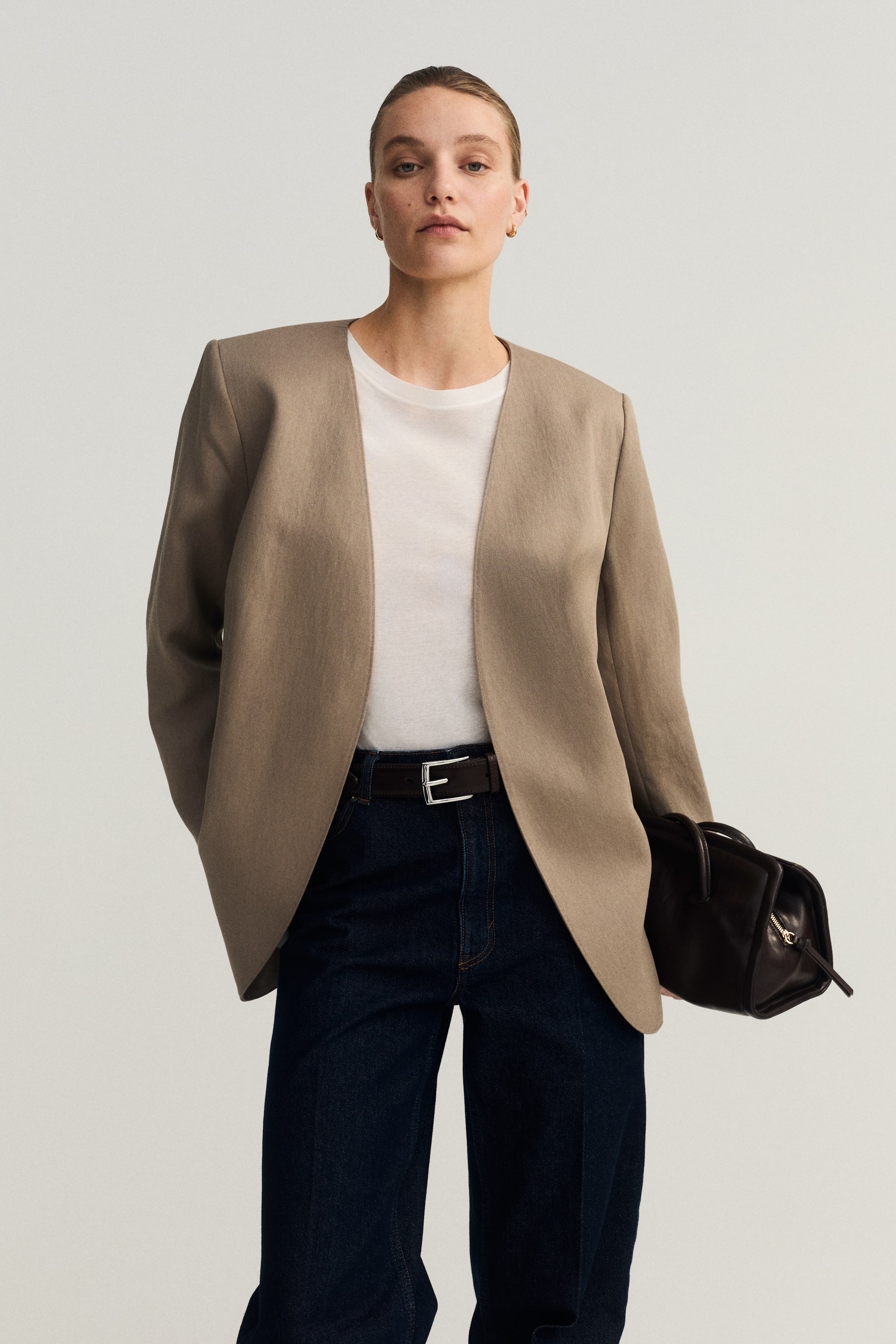 Visa större bild: En taupe kraglös blazer visas framifrån, med en öppen frontdesign, strukturerade axlar och långa ärmar. Den är lagd över en vit t-shirt och mörka jeans.