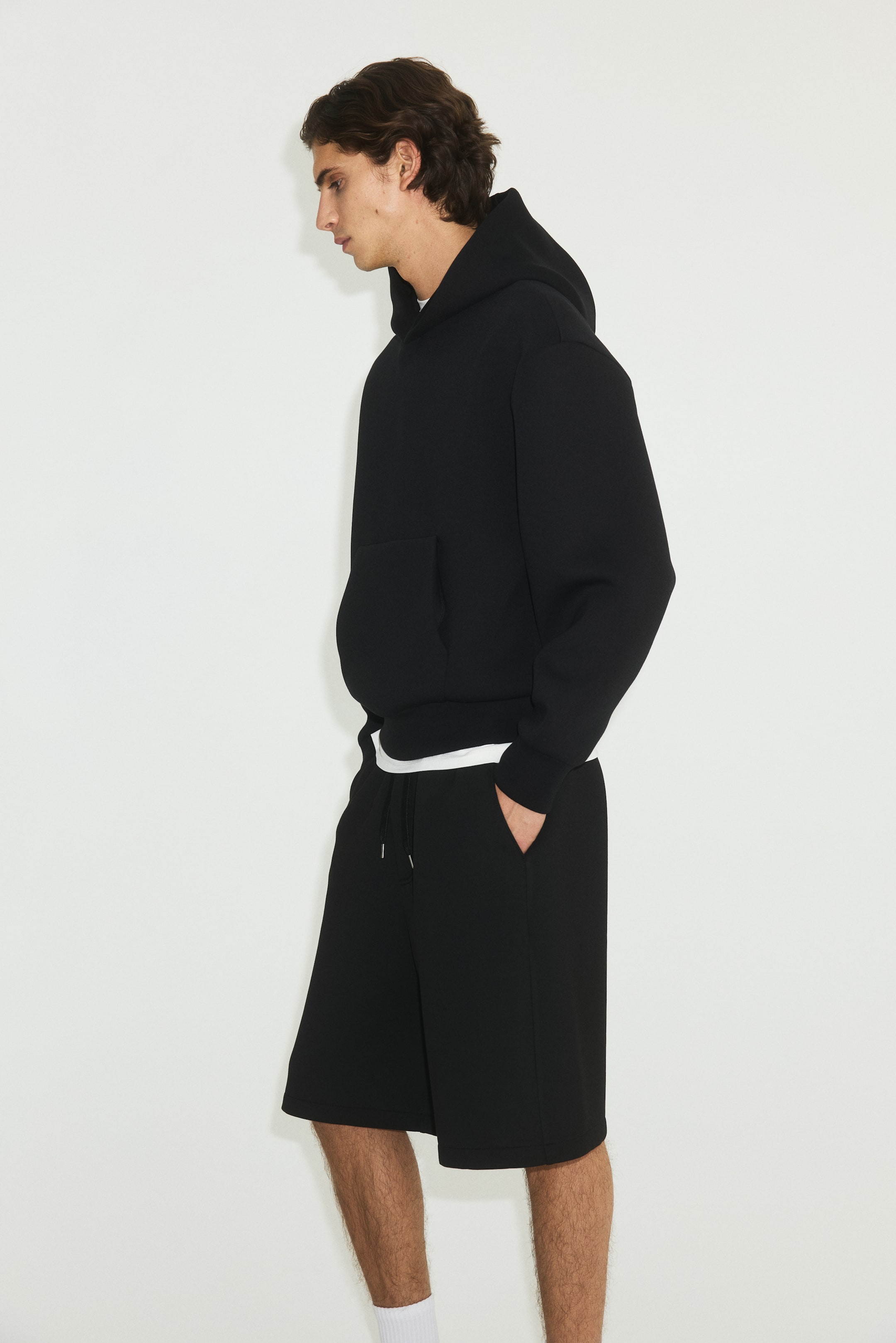 Ampliar la imagen: Una sudadera con capucha negra y shorts negros se presentan desde un ángulo lateral-frontal. La sudadera cuenta con una capucha, mangas largas, un bolsillo canguro al frente y un dobladillo con banda, con una camiseta blanca debajo. Los shorts presentan una cintura con cordón ajustable y bolsillos laterales, terminando por encima de la rodilla y se usan con calcetas blancas.