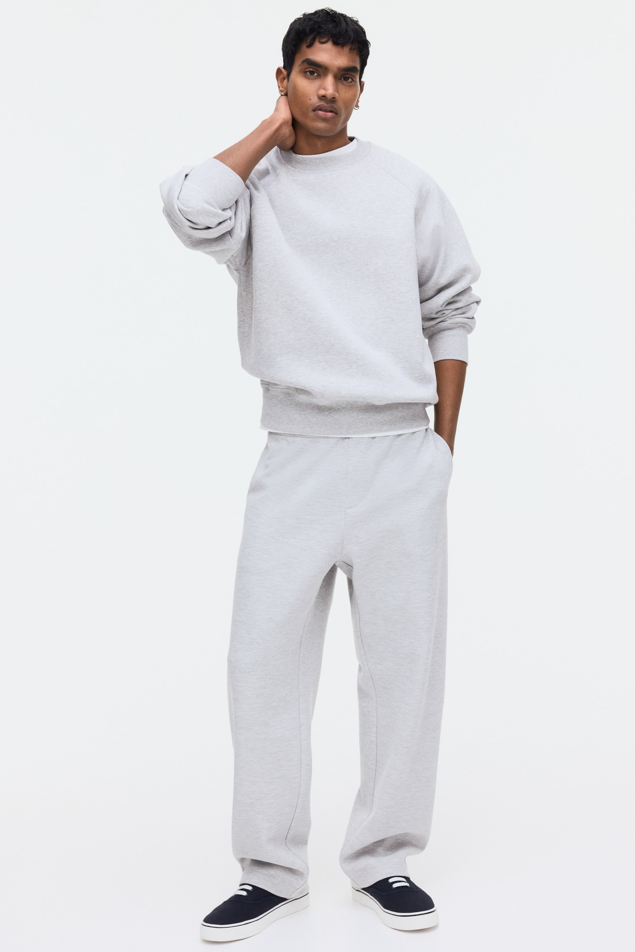 Vezi imaginea mai mare: Pantaloni de trening gri deschis, cu o croială lejeră, talie elastică și craci drepți, sunt purtați de model. Modelul își pune o mână într-un buzunar lateral discret al pantalonilor de trening.