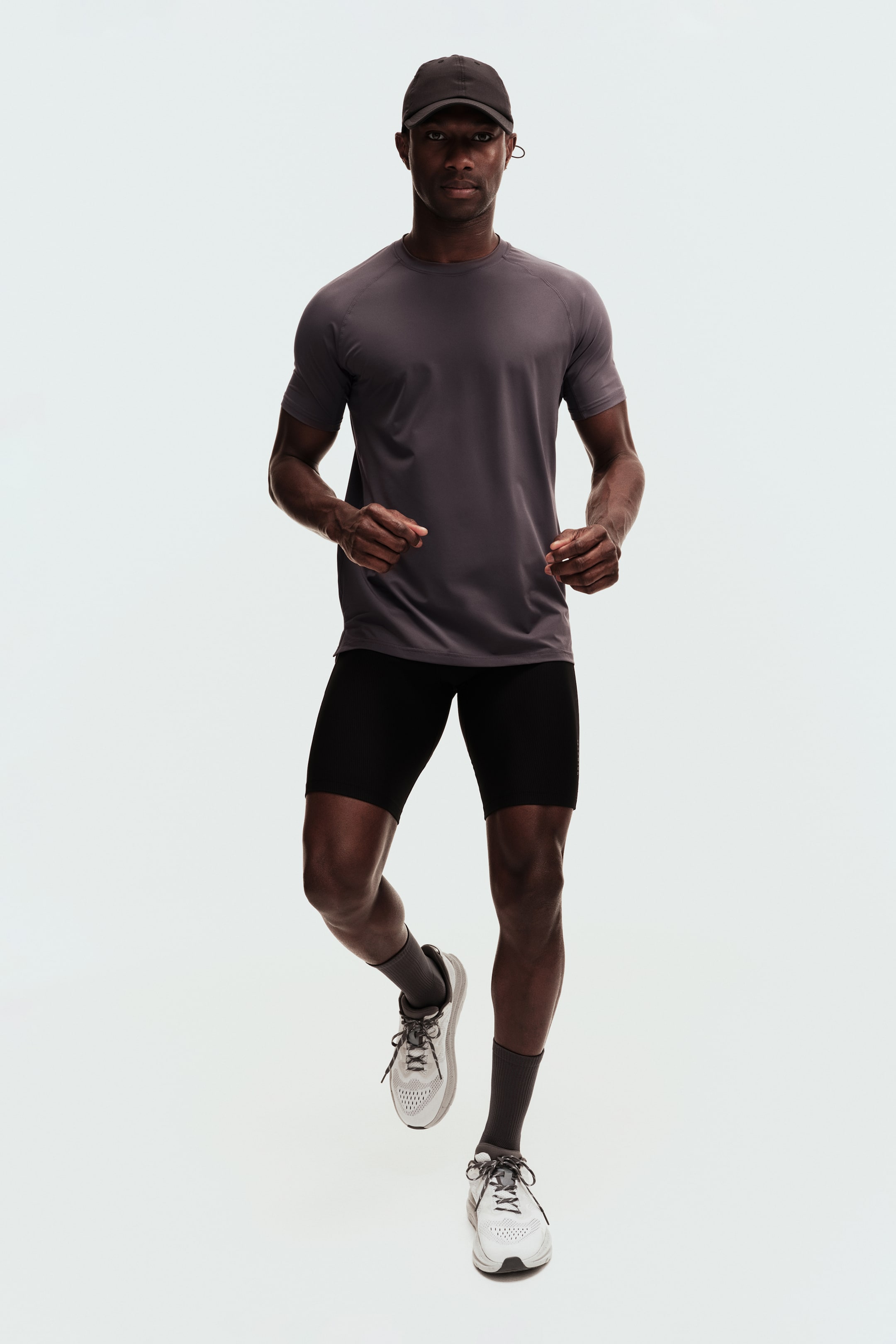 Xem ảnh lớn hơn: Một bộ trang phục thể thao bao gồm áo thun raglan ngắn tay màu xám đậm, quần short ôm sát có gân màu đen, tất thể thao màu xám, giày chạy bộ màu trắng và xám, và mũ bóng chày màu xám đậm, tất cả được trình bày trên một người trong tư thế chạy năng động.