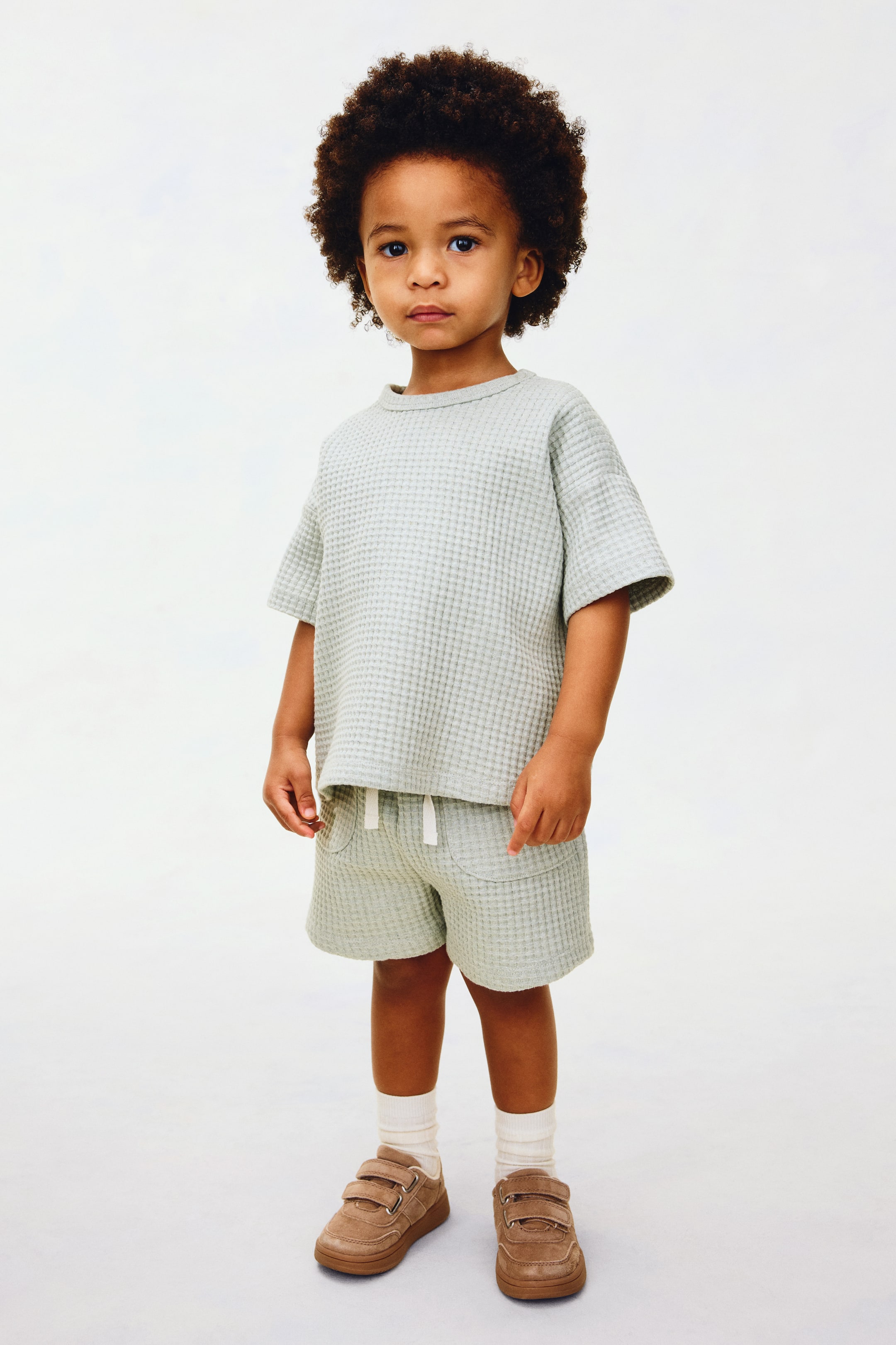 View larger image: A light green waffle-knit short-sleeved t-shirt paired with matching shorts is showcased. The t-shirt features a round neckline, while the shorts come with an elastic waistband, a white drawstring, and two front pockets. Complementing the outfit are brown shoes with two Velcro straps and white ribbed crew socks.
