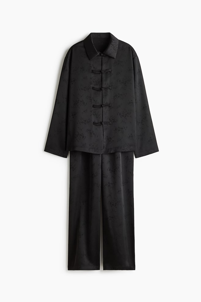 This black jacquard two-piece matching set features a long-sleeved top and matching straight-leg trousers, shown from the front. The top displays a pointed collar and four knotted frog closures down the centre, with both pieces showcasing a subtle, tonal floral pattern on the satin-like fabric. - 1