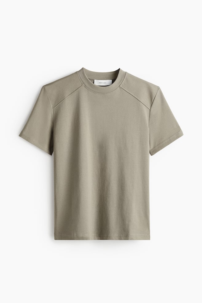 An olive green T-shirt is presented from the FRONT, featuring a ribbed crew neck, short sleeves, and a double-layered yoke with angled shoulder seams. A white brand label is visible at the inner back neck. - 1