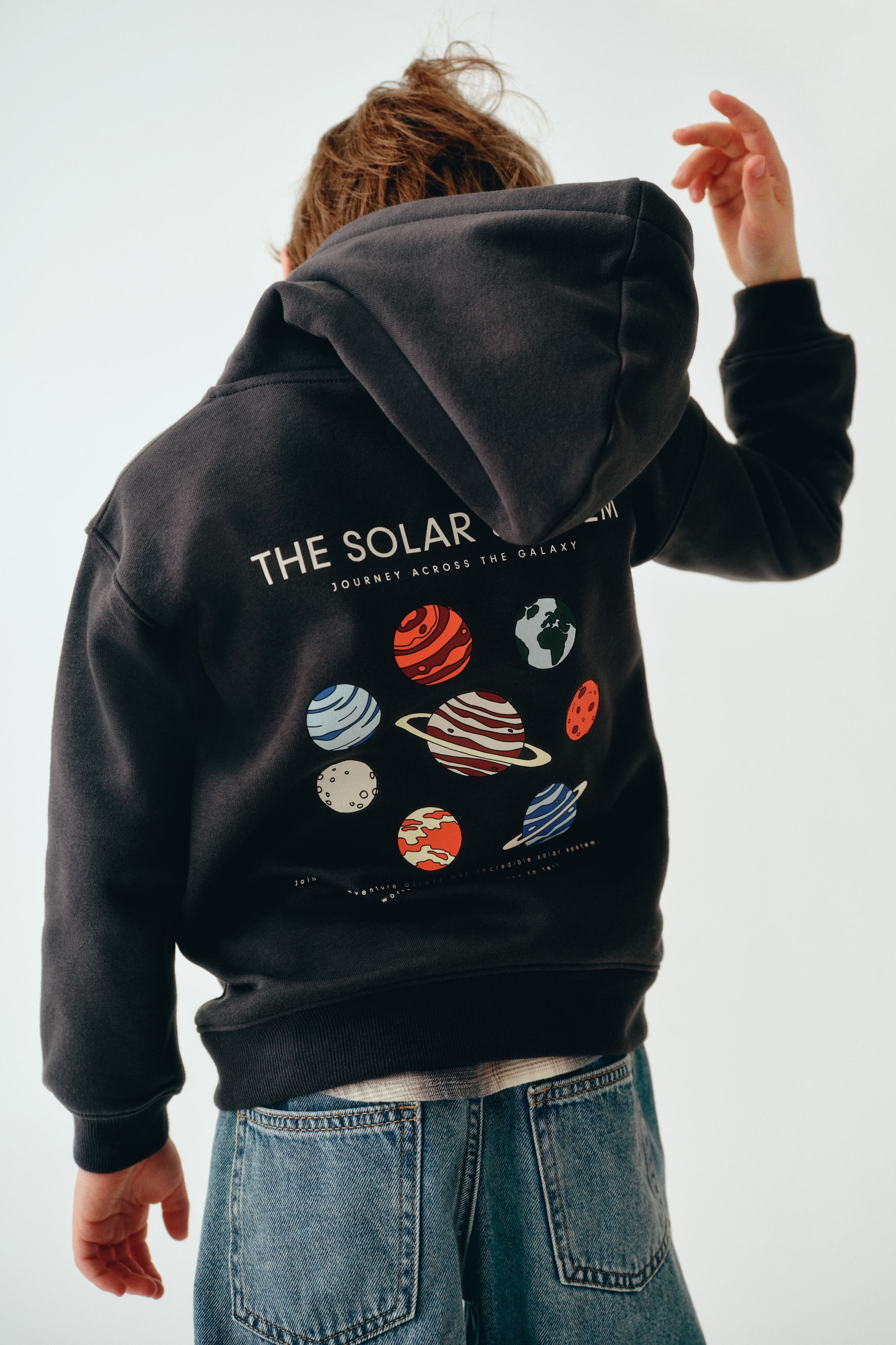 Grotere afbeelding bekijken: Een donkergrijze hoodie met losse pasvorm wordt van achteren getoond, met een grote grafische print van het zonnestelsel met kleurrijke planeten en witte tekst met de woorden "THE SOLAR SYSTEM". De hoodie is ontworpen met verlaagde schouders, een nonchalante capuchon en brede geribde boorden aan de mouwen en onderzoom.
