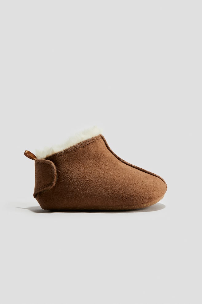 Une seule pantoufle de style bottine marron est positionnée en vue latérale, avec un extérieur doux semblable à du daim, une doublure en shearling de couleur crème visible autour de l'ouverture, et une petite languette marron s'étendant depuis le talon. - 1
