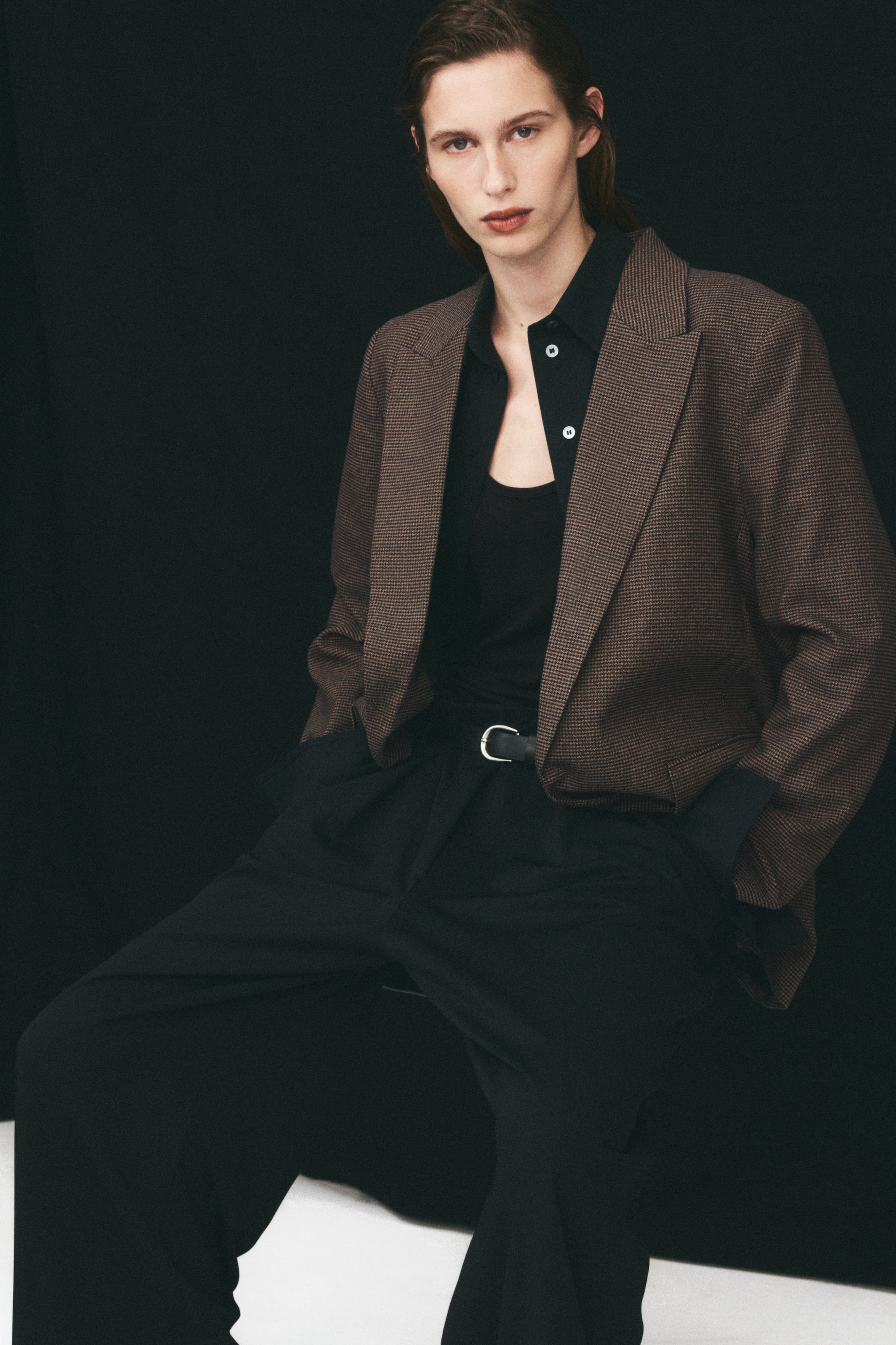 Agrandir l'image: Un blazer long et oversize en tissu tissé à motif brun et noir est porté par le mannequin. Le blazer présente des revers à pointe et se porte ouvert sur une chemise noire boutonnée et un pantalon noir.