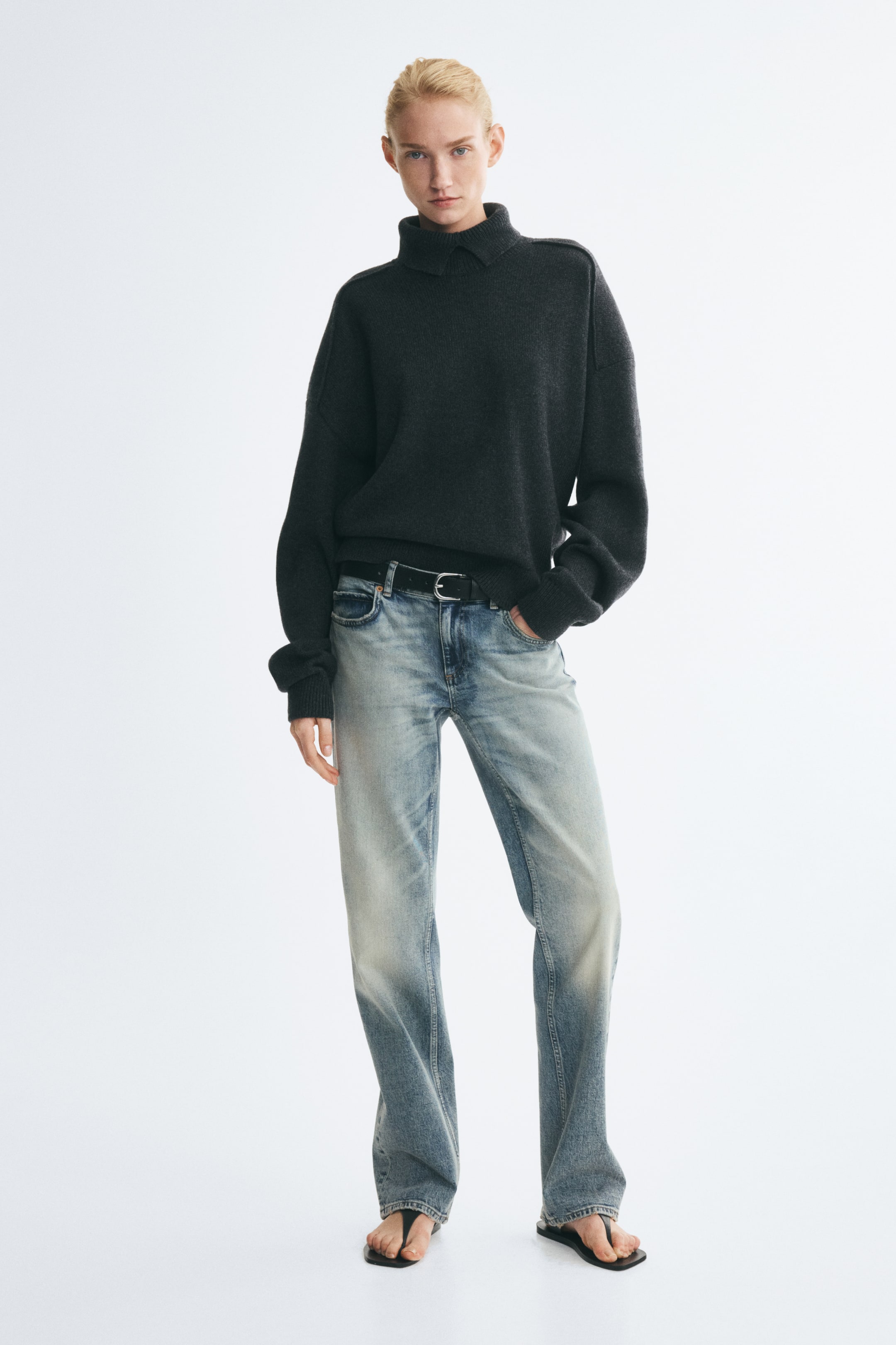 Se større billede: Lyse, straight-leg denimjeans med lav talje og falmede detaljer bæres af modellen, som står med den ene hånd hvilende i en forlomme. Jeansene er stylet med et sort læderbælte, en mørkegrå sweater med høj hals og flade sorte sandaler.