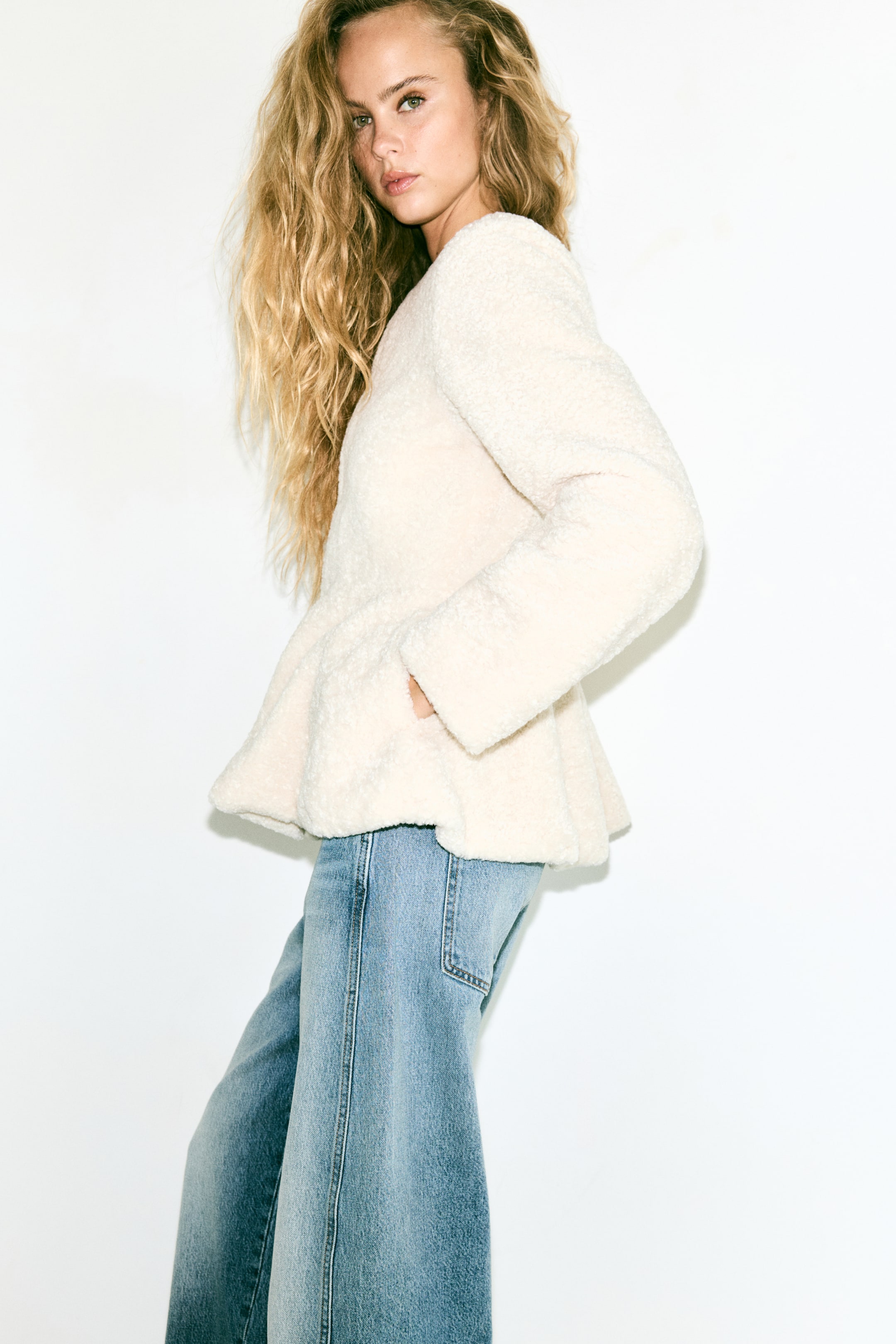Se større billede: En cremefarvet pilefleecejakke med rund halsudskæring, lange ærmer og en udsvinget peplum bæres af modellen. Modellen hviler den ene hånd i jakken side lomme, mens hun har blå jeans på.