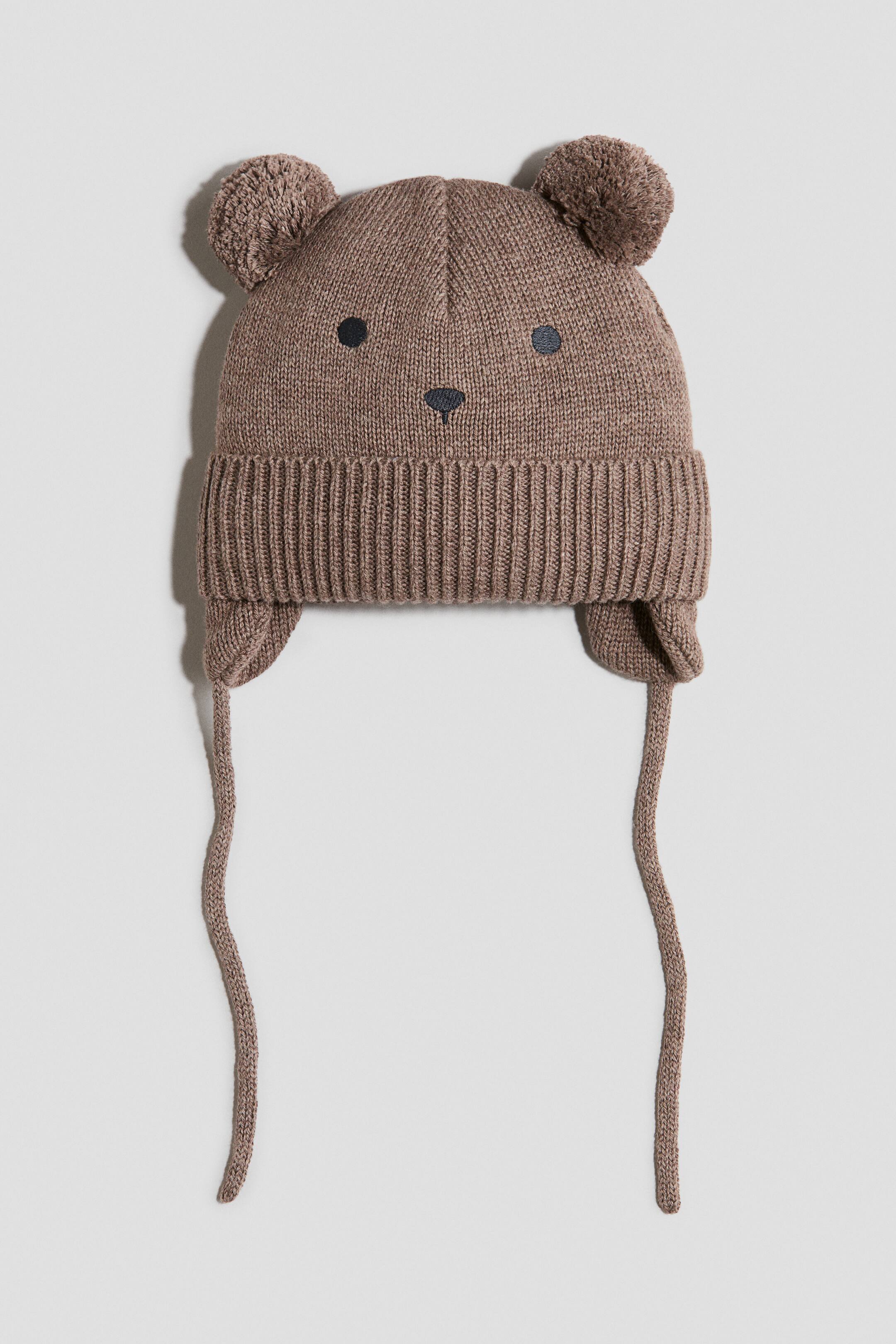 View larger image: The brown knitted hat is shown from the front, featuring two rounded pom-pom ears on top and an embroidered face with black circular eyes and a small triangular nose. It features a ribbed brim, ear flaps, and braided ties hanging from each flap.