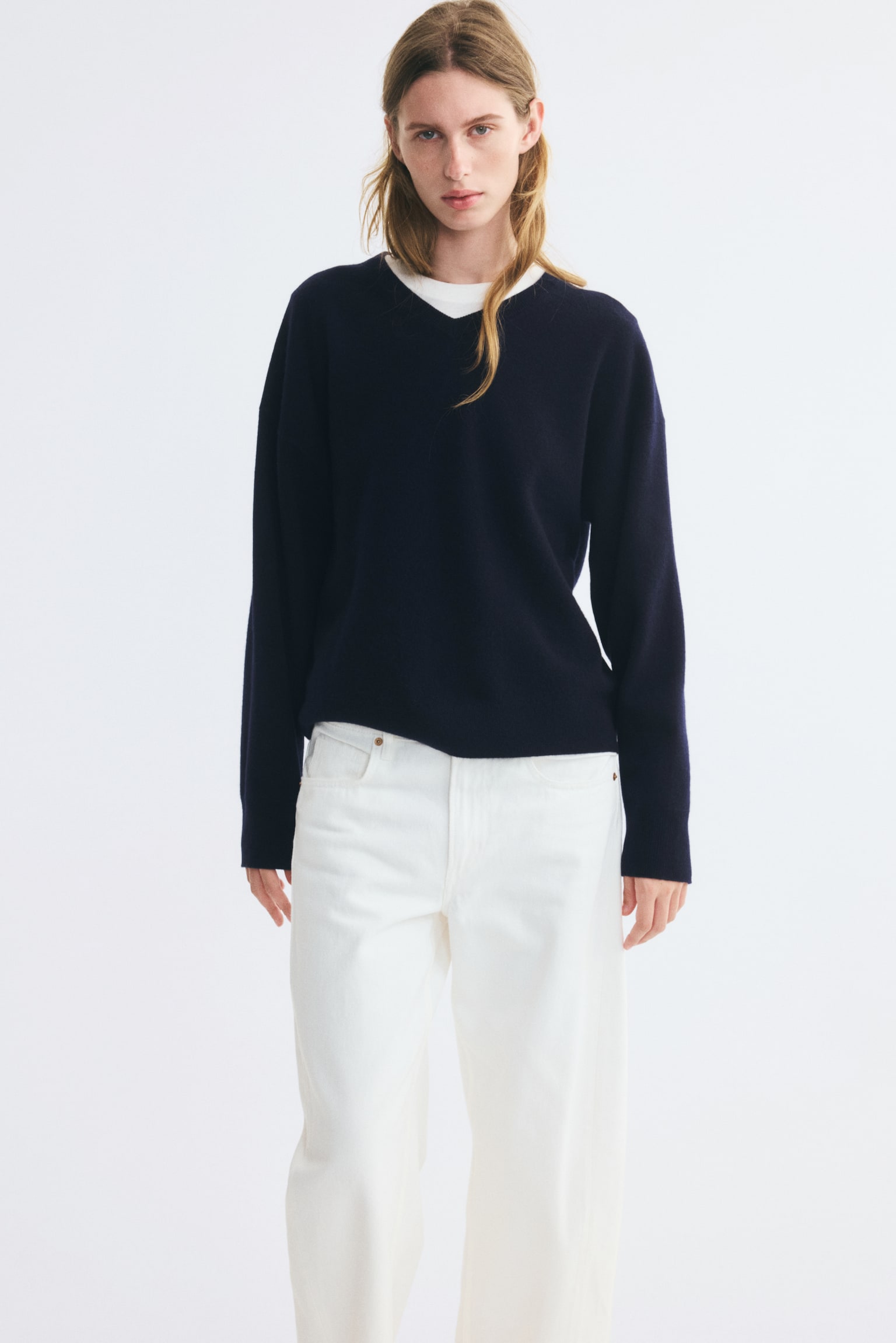 A dark navy, fine-knit wool sweater featuring a ribbed V-neck, dropped shoulders, and ribbed cuffs is being worn by the model. The oversized sweater is layered over a white crew-neck T-shirt and paired with white trousers. - 1
