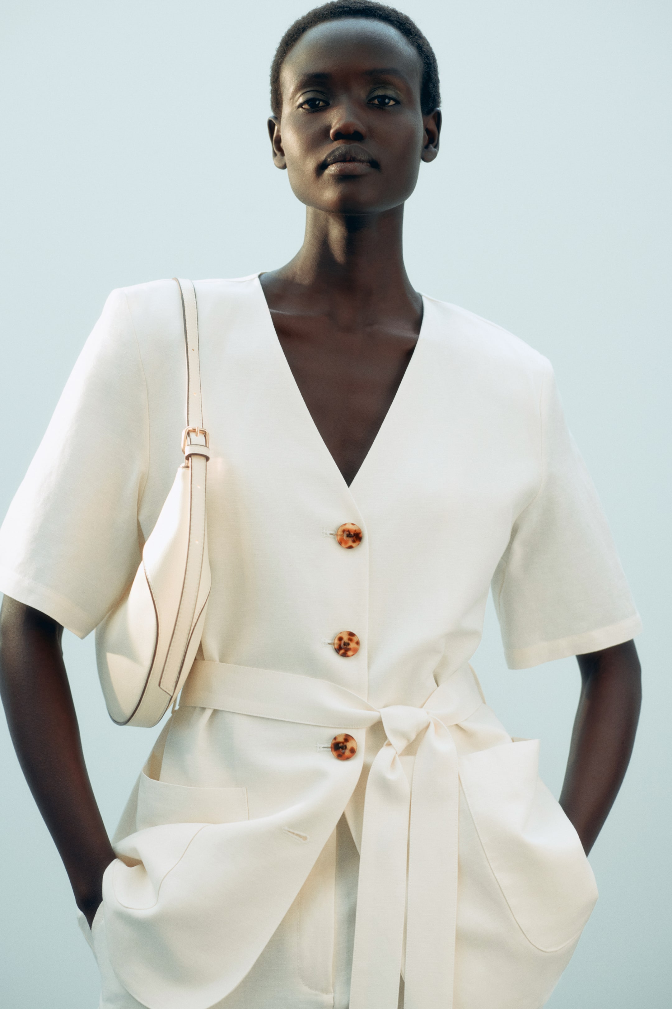 View larger image: An off-white blazer in an airy linen blend, featuring a V-neckline, short sleeves and tortoiseshell buttons down the front, is being worn by the model. A tie belt is being fastened at the waist, the model is placing their hands in the patch front pockets, while a shoulder bag rests on the blazer.