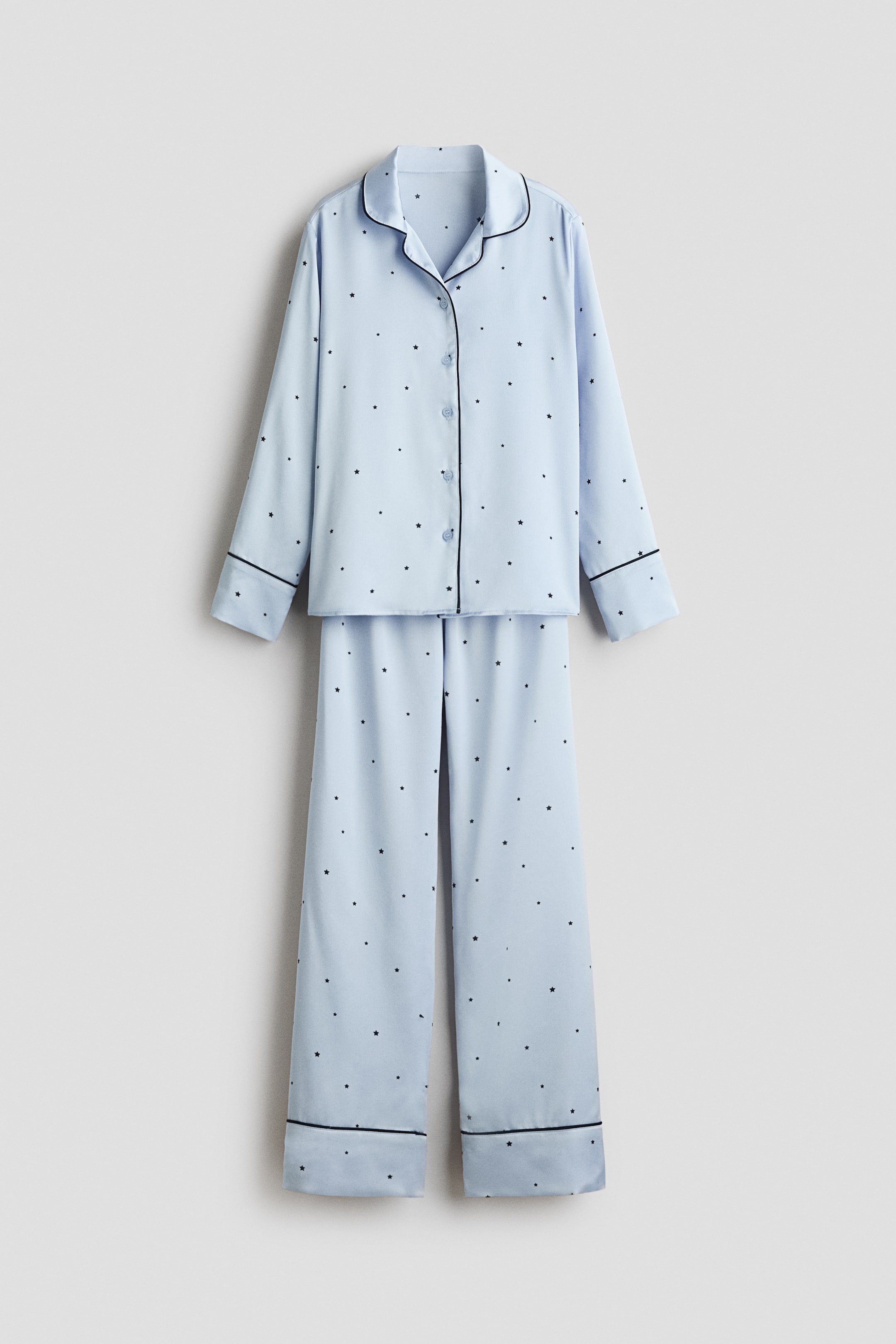 View larger image: A light blue satin pyjama set is shown from the front, featuring a matching long-sleeve shirt and straight-leg pants decorated with a small black star pattern. The shirt features a resort collar, front button closures, and dark piping along the collar and cuffs, while the pants feature matching piping at the hems.