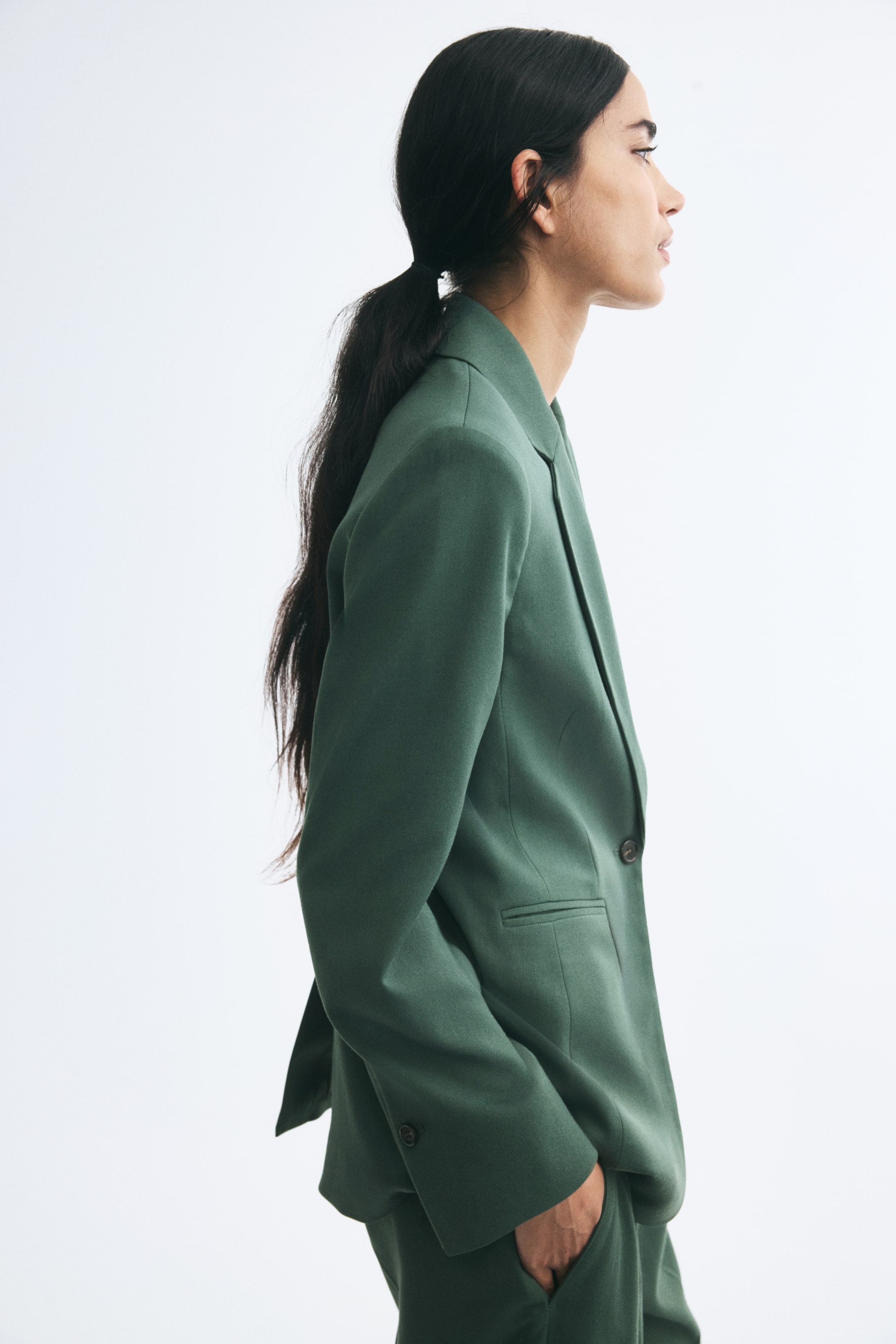 View larger image: A green, single-breasted blazer featuring notch lapels, a jetted pocket, and button cuffs is worn by the model. The blazer is paired with matching green trousers, with the model standing in profile and placing a hand in the trouser pocket.