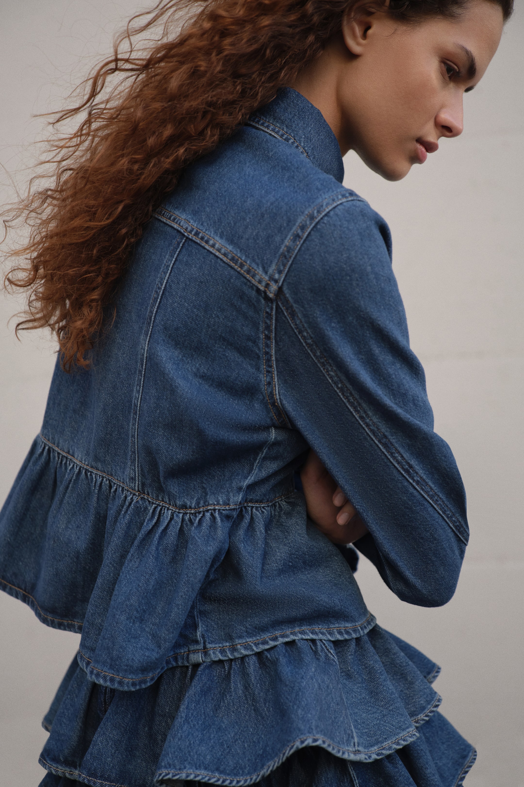 View larger image: A denim jacket featuring a stand-up collar and a ruffled peplum hem is showcased from the back and side. It is paired with a coordinating tiered denim skirt, highlighting multiple ruffled panels.