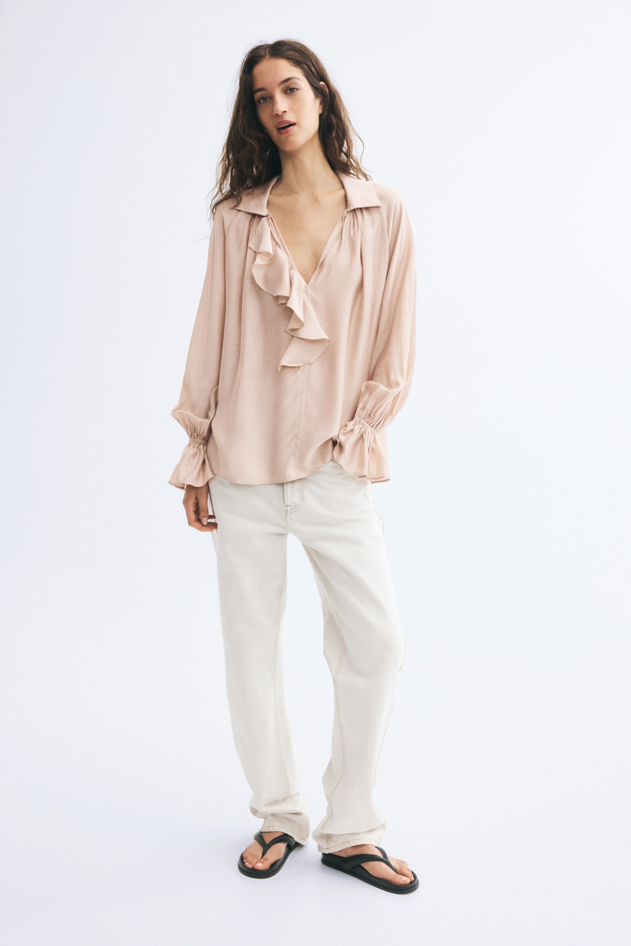 Xem ảnh lớn hơn: Người mẫu đang mặc một chiếc áo blouse màu be hồng nhạt, có cổ áo, cổ chữ V sâu, chi tiết bèo xếp tầng phía trước và tay áo dài phồng với cổ tay bèo. Áo được phối dáng suông cùng quần màu trắng ngà.