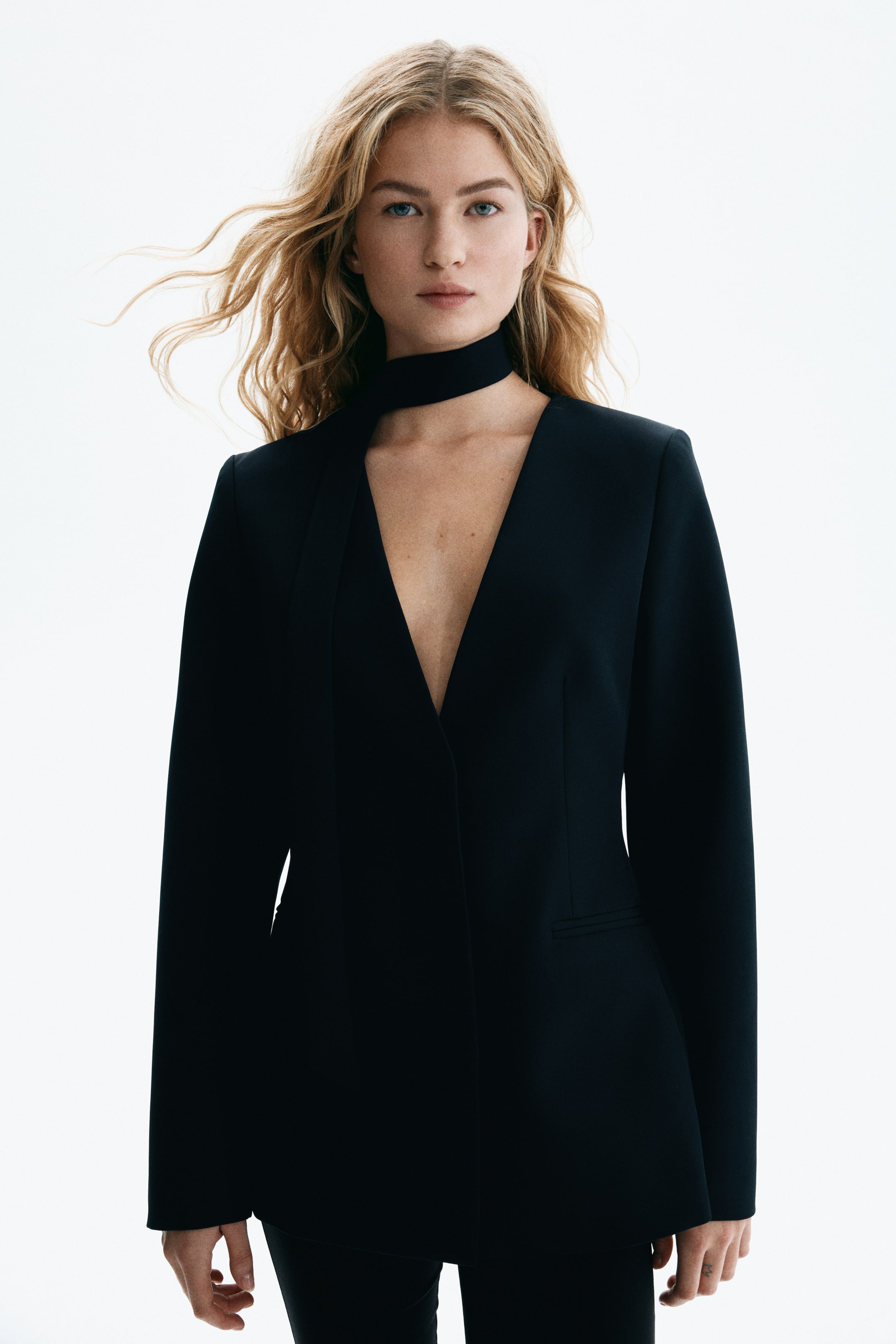 View larger image: A black blazer features a deep V-neckline, structured shoulders, and a front flap pocket. A black choker-style scarf complements the blazer's open neckline.