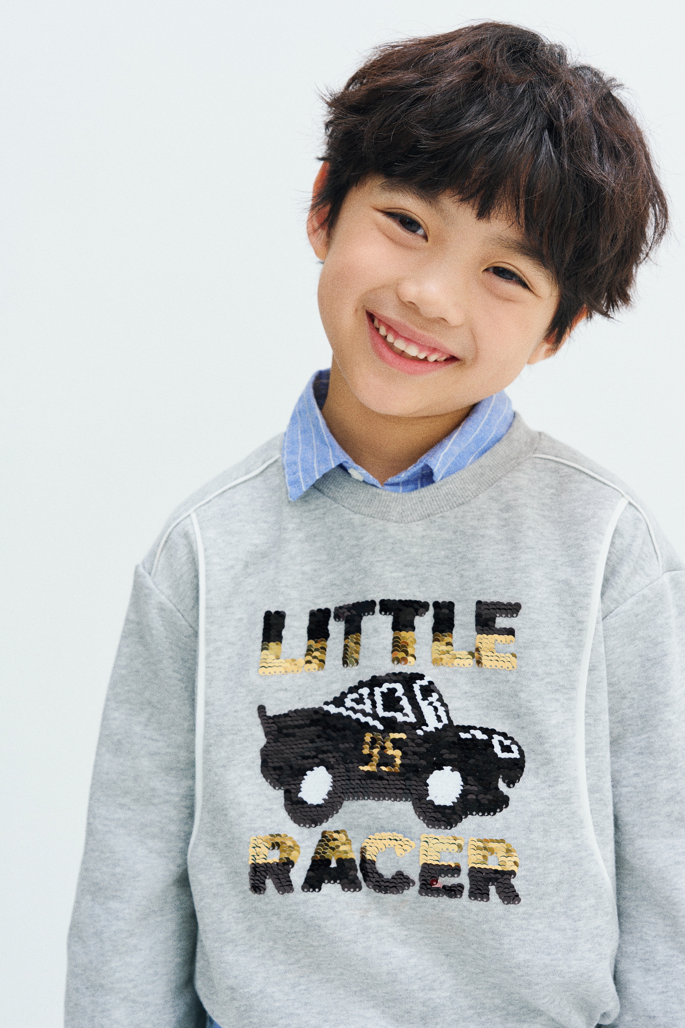A light grey sweatshirt is shown from the front, featuring a ribbed crew neck, ribbed cuffs, and a ribbed hem. The sweatshirt highlights curved front seams with white piping and a central sequinned graphic of a red race car between the words "LITTLE RACER" in blue, red, and black.