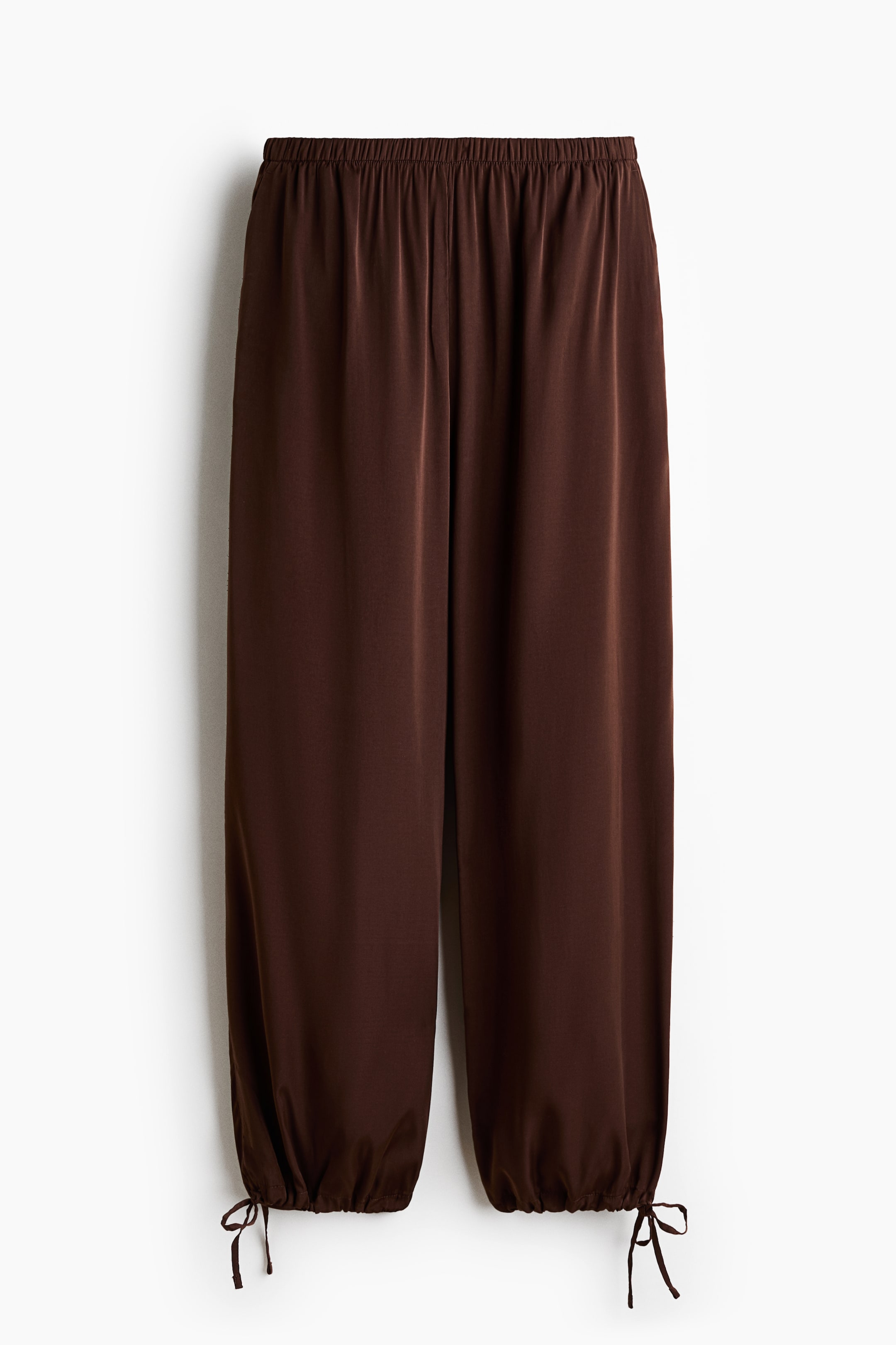 View larger image: Brown pants are showcased from the front, featuring an elastic waistband and a smooth, flowing fabric. The full-length legs end in gathered cuffs with visible tie details.