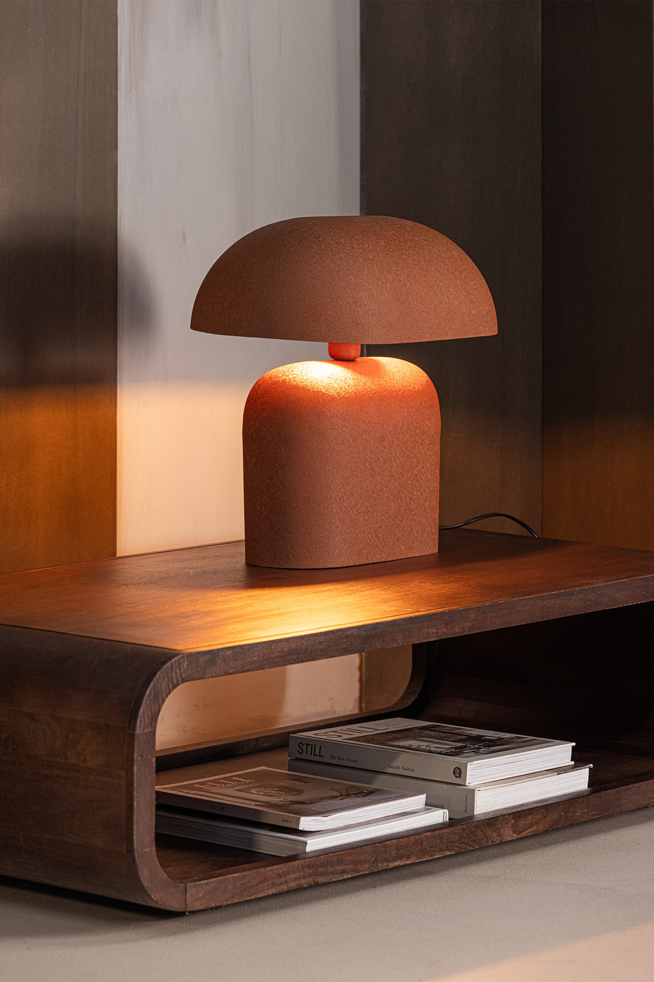Größeres Bild ansehen: Eine Terrakotta-Tischlampe erleuchtet den Raum und besticht durch einen kompakten, abgerundeten Sockel sowie einen halbkugelförmigen Schirm, der durch einen kleineren, helleren orangefarbenen Zylinder verbunden ist. Die Lampe präsentiert sich mit einer strukturierten, matten Oberfläche.