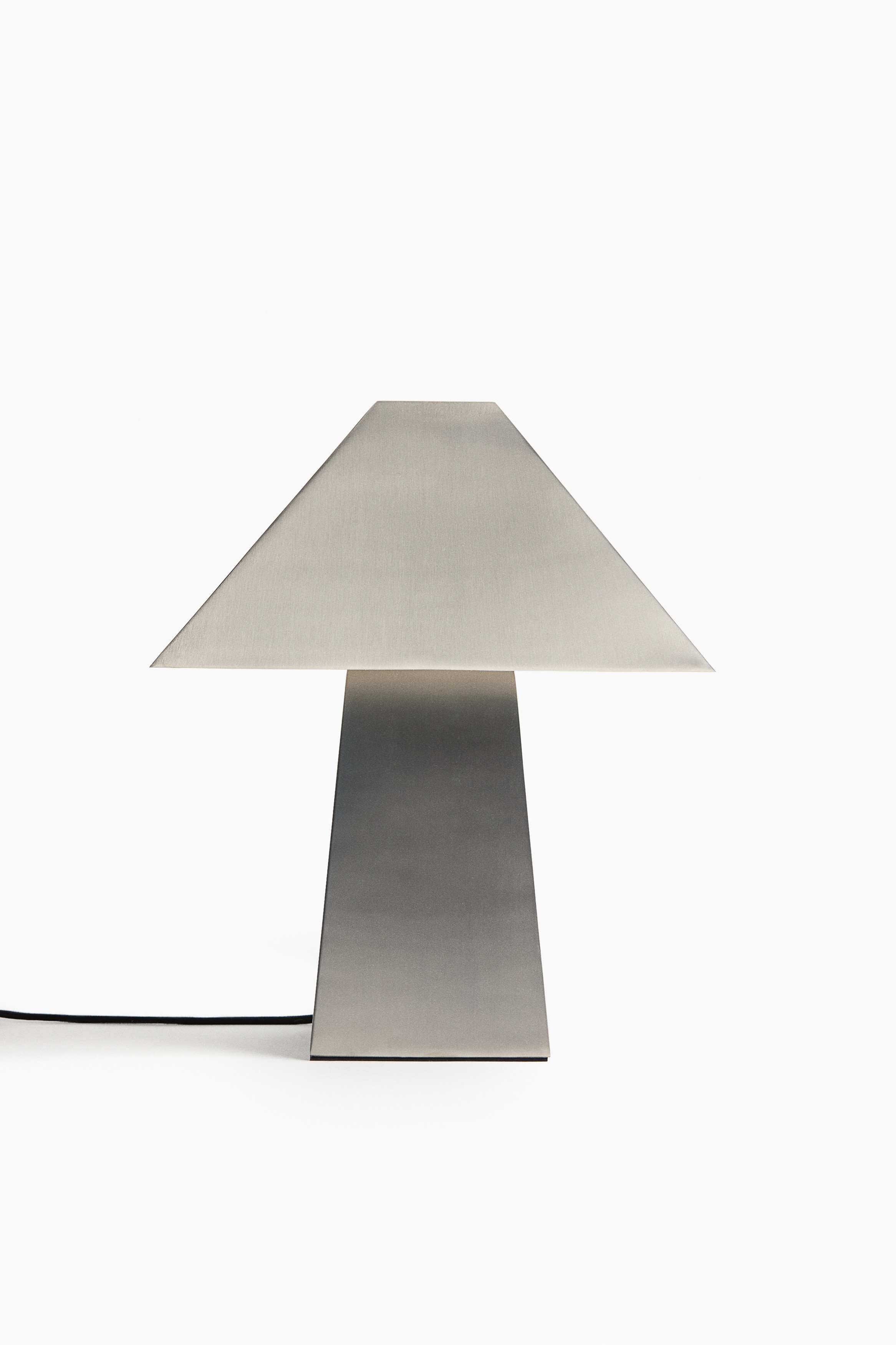 A front-facing metal table lamp featuring a geometric design with a trapezoidal shade and a tapering rectangular base is standing illuminated. A warm light is glowing from beneath the shade, casting a soft reflection onto the brushed metal surface of the base.