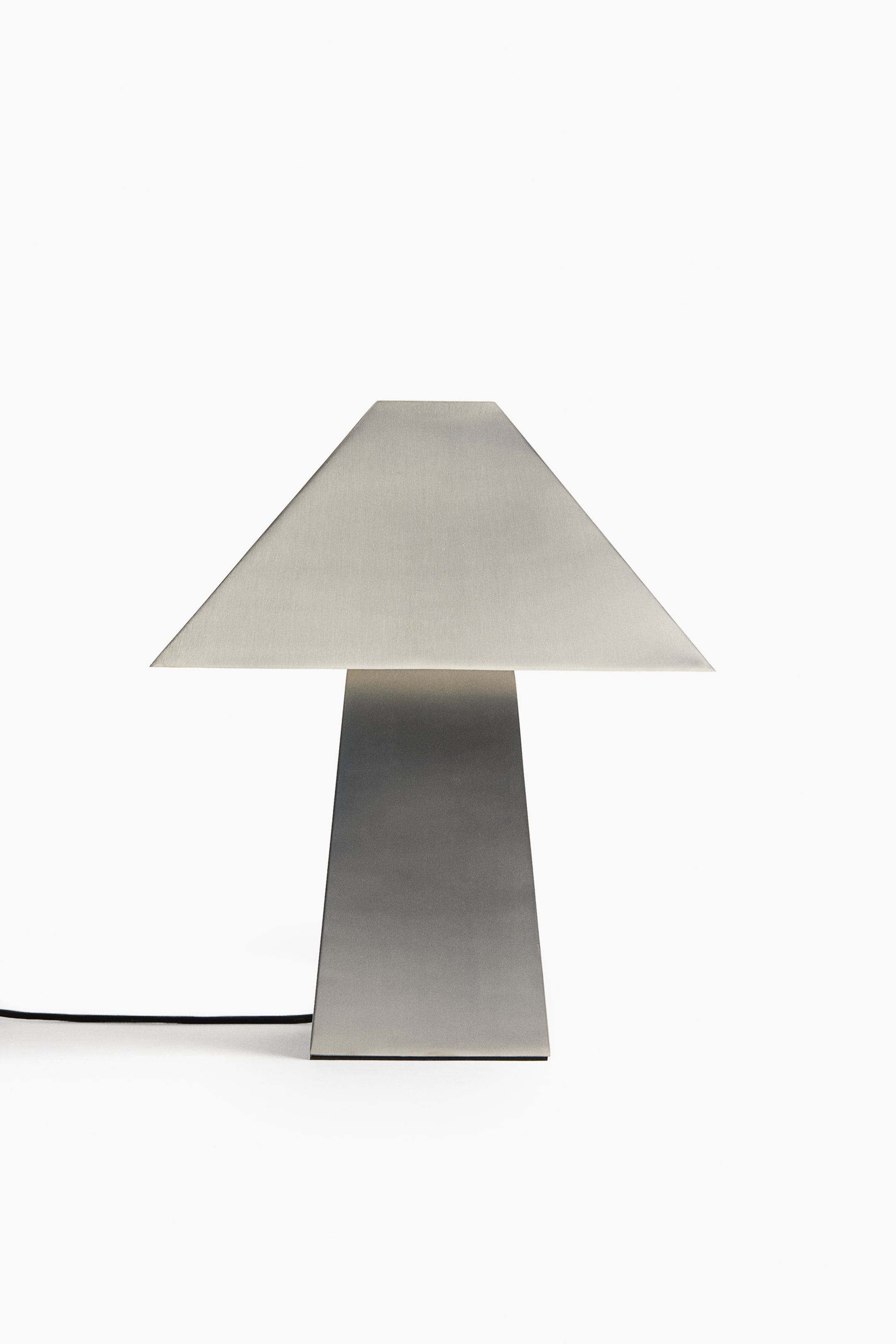 Se større billede: En metallisk bordlampe står vendt mod fronten og fremhæver en børstet ståloverflade. Den har en trapezformet skærm, der hviler på en konisk, rektangulær søjlebase med en sort ledning, der løber ud fra nederste venstre side.
