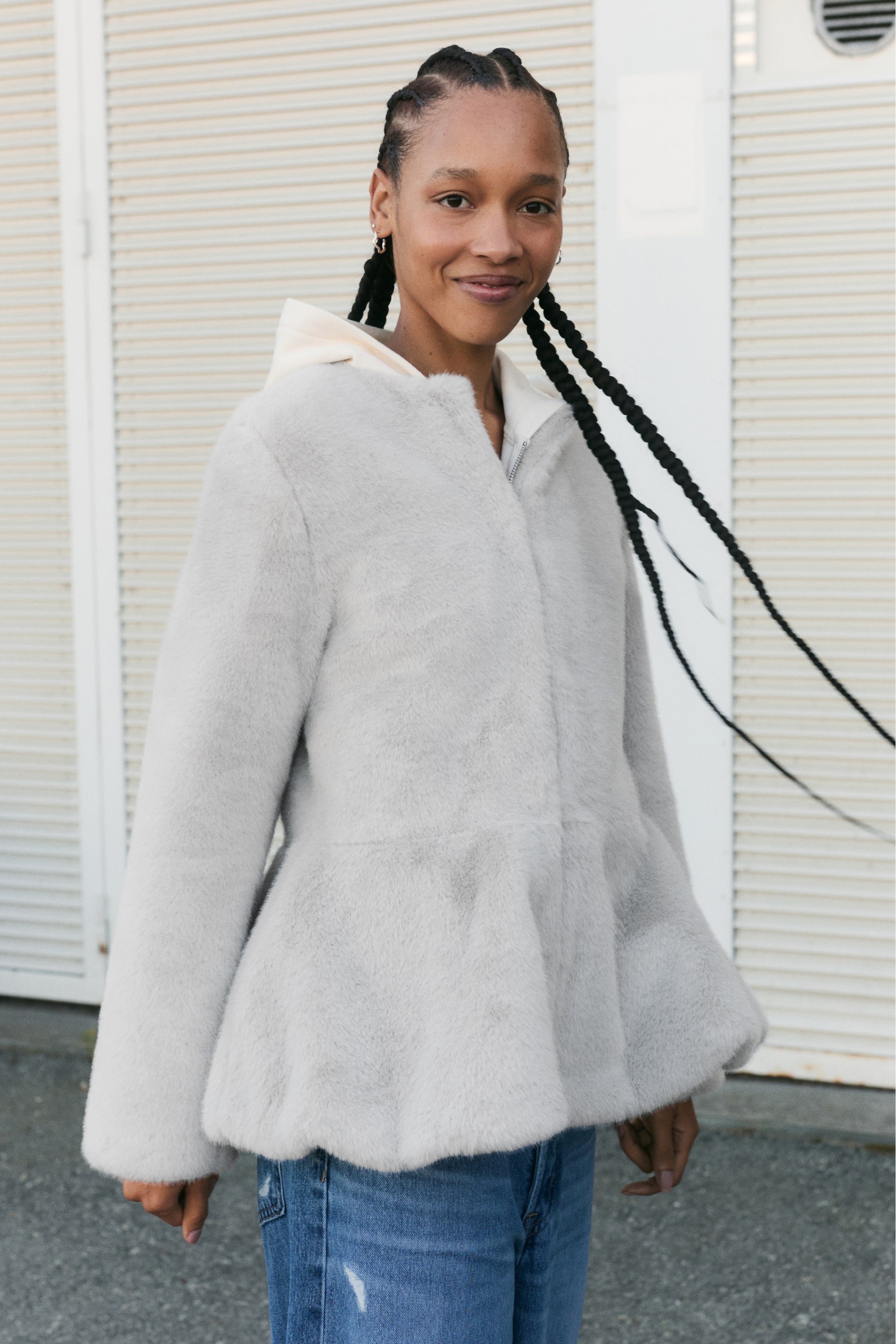 Größeres Bild anzeigen: Ein hellgraues Jacket aus weichem Pile-Fleece wird vom Model getragen und verfügt über einen runden Ausschnitt, lange Ärmel und einen ausgestellten Peplum-Saum. Das Jacket wird über einem cremefarbenen Hoodie getragen und mit blauen Jeans kombiniert.