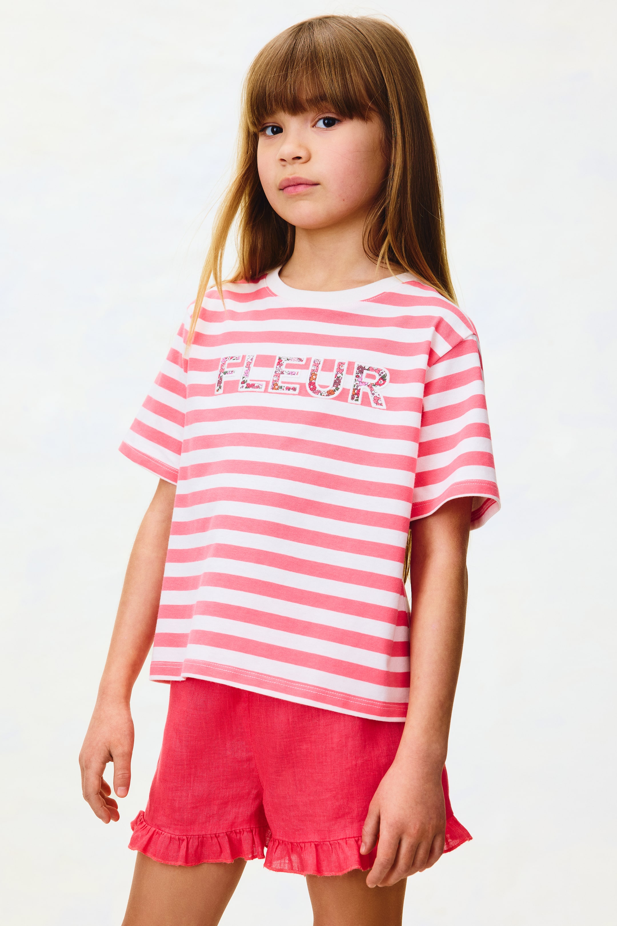 Se større billede: En hvid og lyserød vandretstribet t-shirt viser ordet 'FLEUR' i et blomsterprint hen over brystet. Matchende lyserøde shorts har flæsekantede sømme.