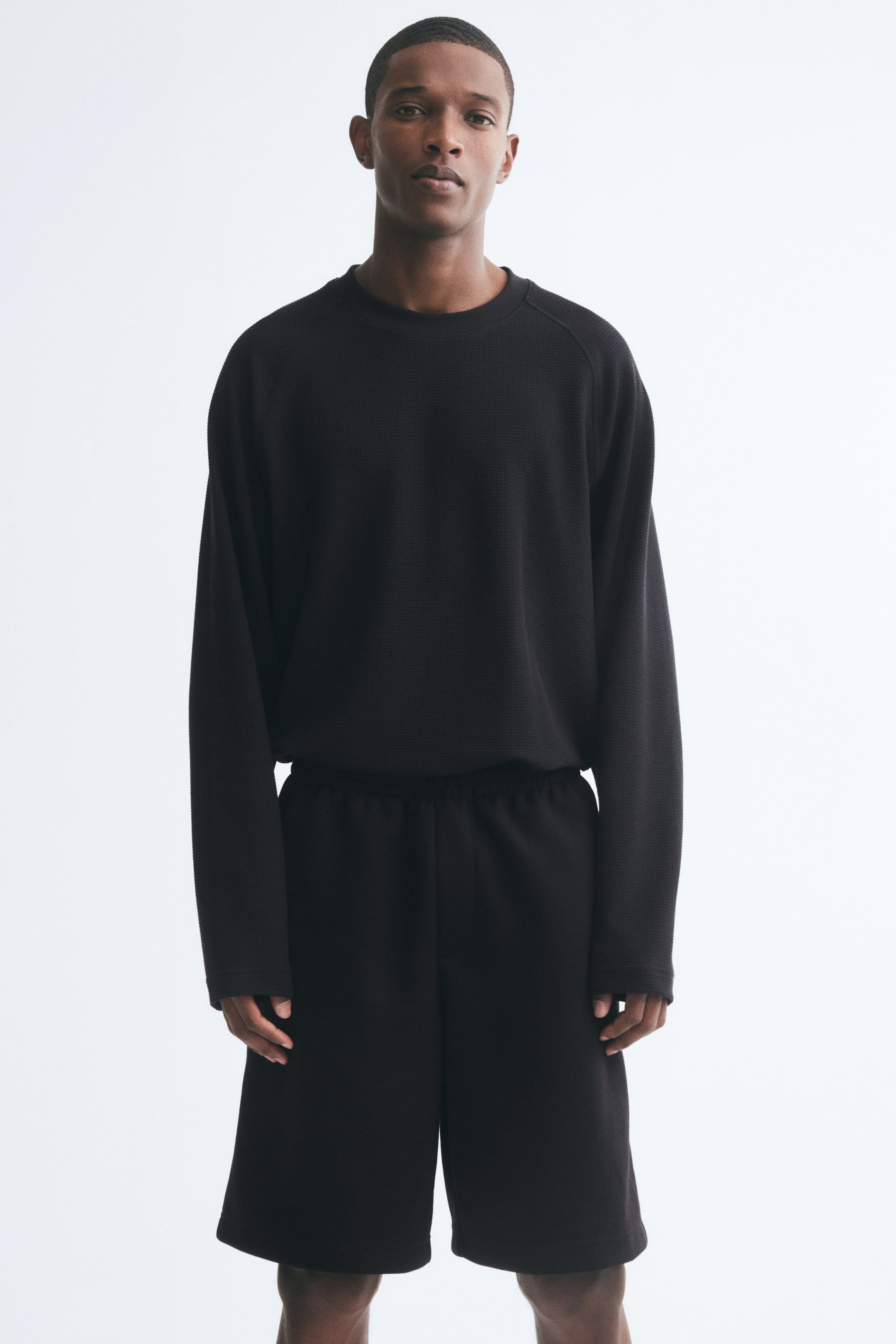 Grösseres Bild ansehen: Schwarze Sweatshorts in Kniehöhe mit lockerer Passform, elastischem Bund und angedeutetem Hosenschlitz werden vom Model getragen. Die Shorts sind mit einem passenden schwarzen, strukturierten Langarm-Rundhalstop kombiniert.