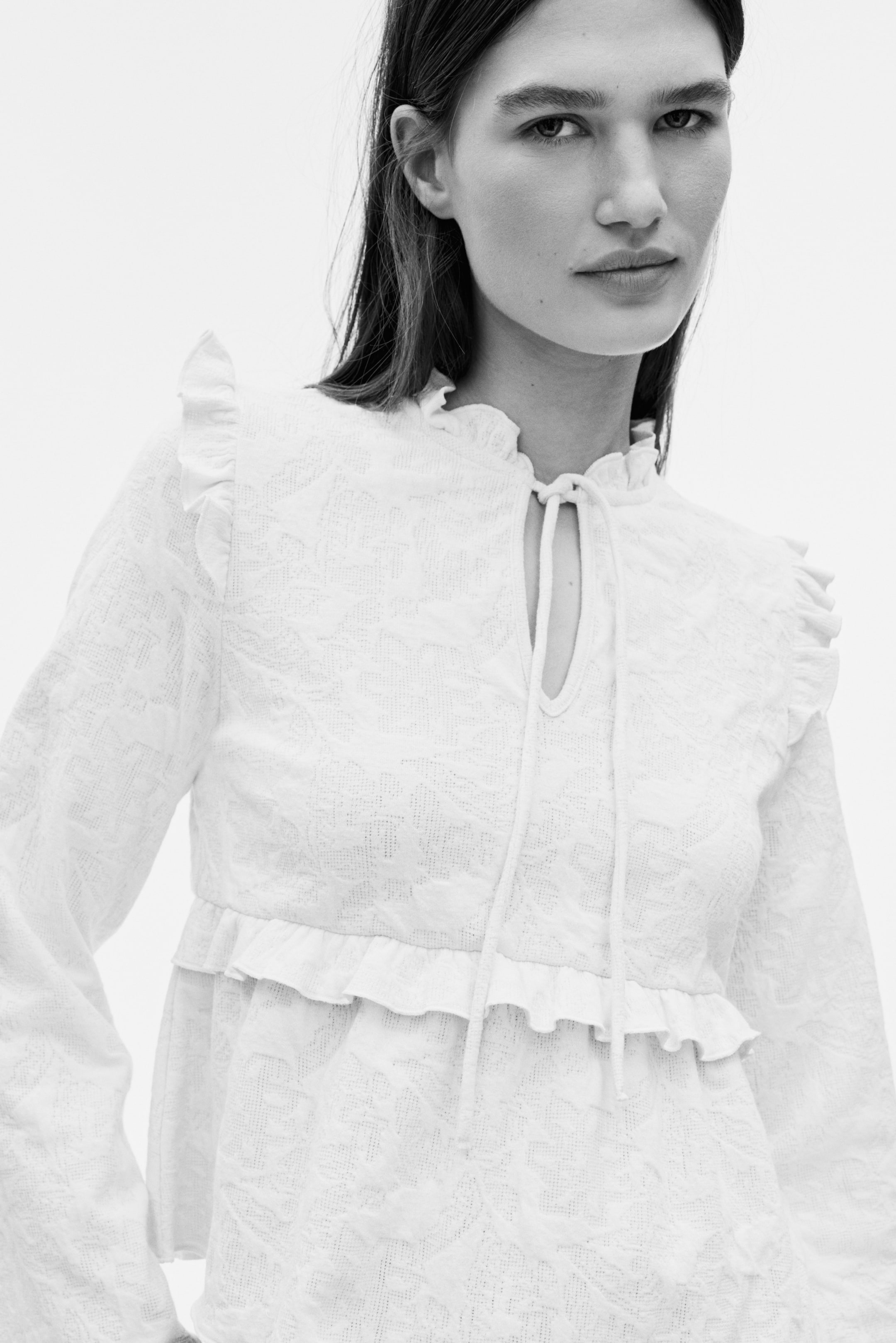 View larger image: White long-sleeved blouse shown facing forward, featuring an embossed floral pattern. It has a ruffled high collar, a keyhole neckline with tie strings, ruffled shoulder details, and a ruffled peplum hem.