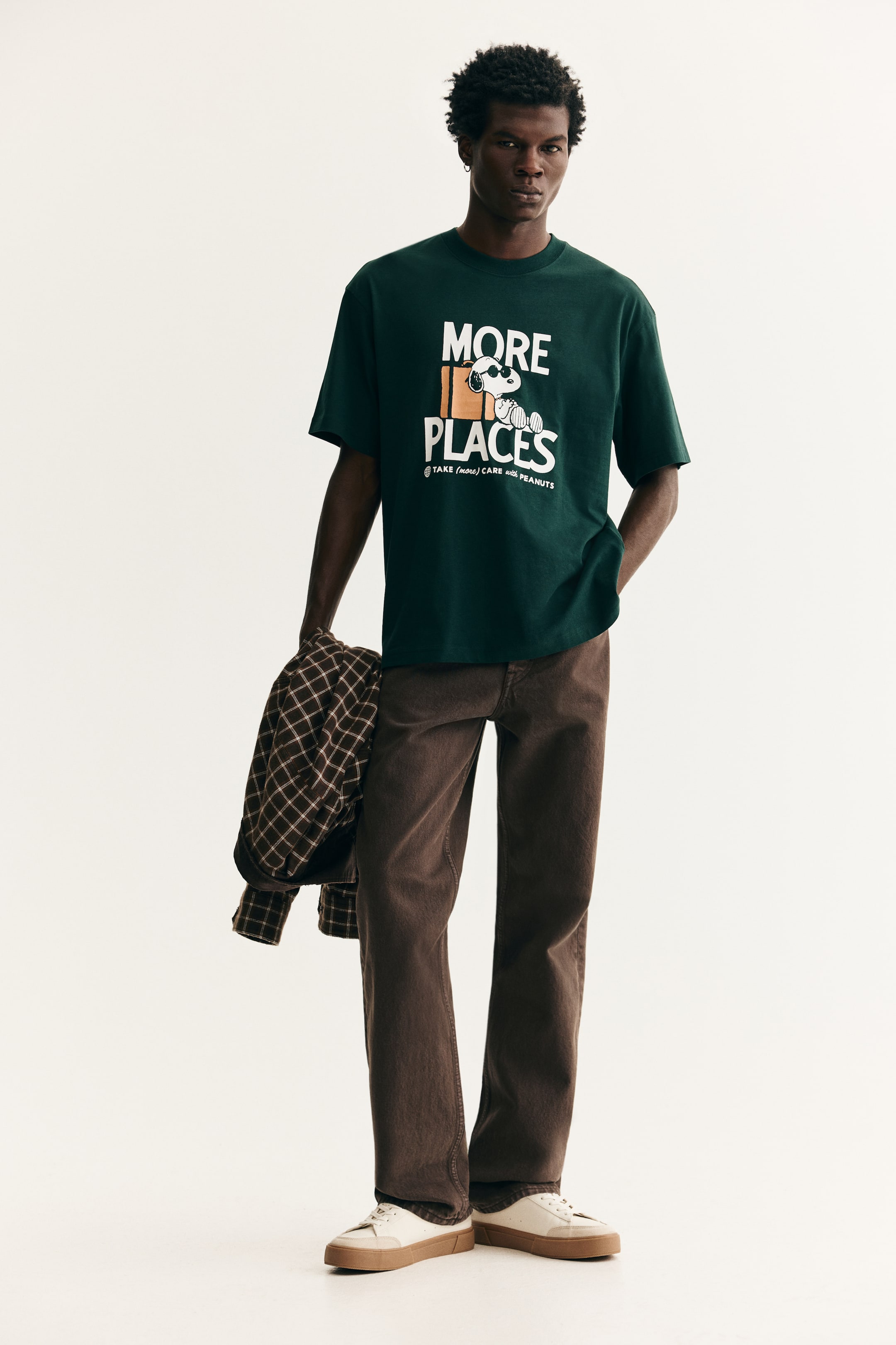 View larger image: A dark green crewneck t-shirt is being shown front-facing, featuring a white graphic print on the chest. The graphic includes the stacked text "MORE PLACES" with an image of Snoopy wearing sunglasses and holding a suitcase between the words, and smaller text "TAKE (MORE) CARE WITH PEANUTS" below.