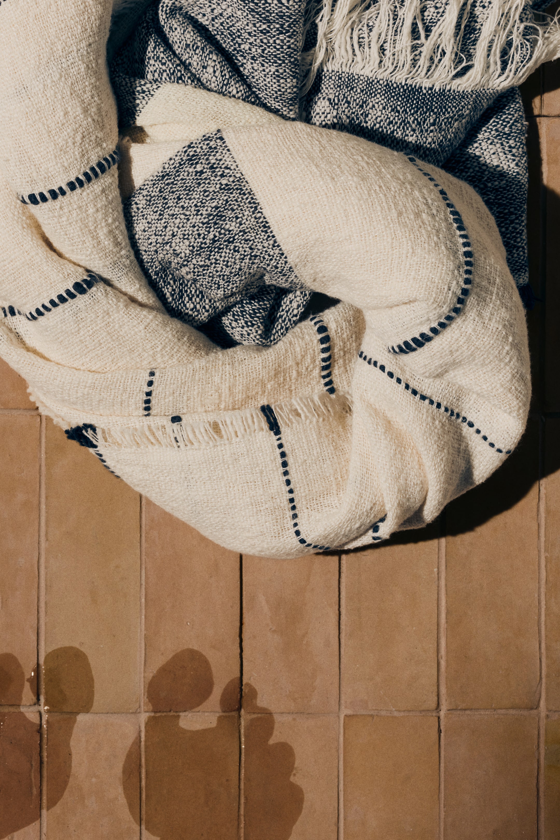 View larger image: A woven blanket, viewed from an overhead angle, is draped in a loose circular shape. It features cream-coloured sections with dark dashed lines forming a grid pattern, and dark, speckled sections with fringes appearing along some edges.