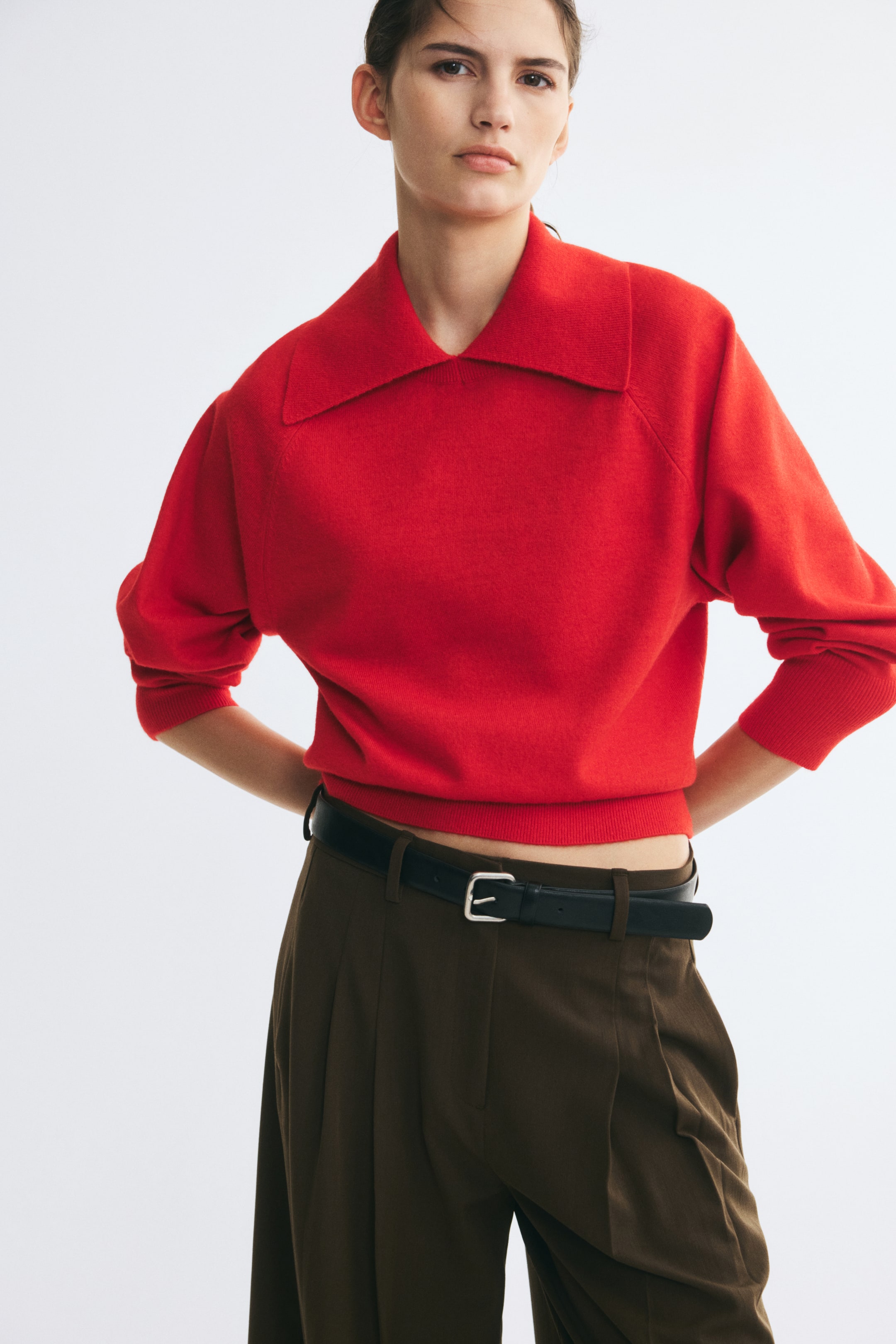 Grösseres Bild ansehen: Ein roter, weich gestrickter Oversize-Pullover mit grossem, flachem Kragen, langen Raglanärmeln sowie gerippten Bündchen und Saum wird vom Model getragen. Der Pullover ist mit braunen Bundfaltenhosen und einem schwarzen Gürtel kombiniert.