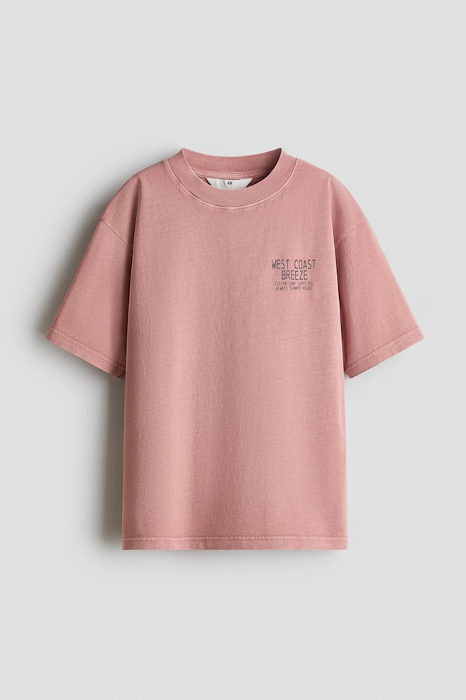 Un t-shirt rose à coupe ample en jersey de coton doux se présente de face. Il est doté d'un col rond bordé de côtes, d'épaules légèrement tombantes, et d'un motif texte noir imprimé sur la partie supérieure droite de la poitrine. - 1
