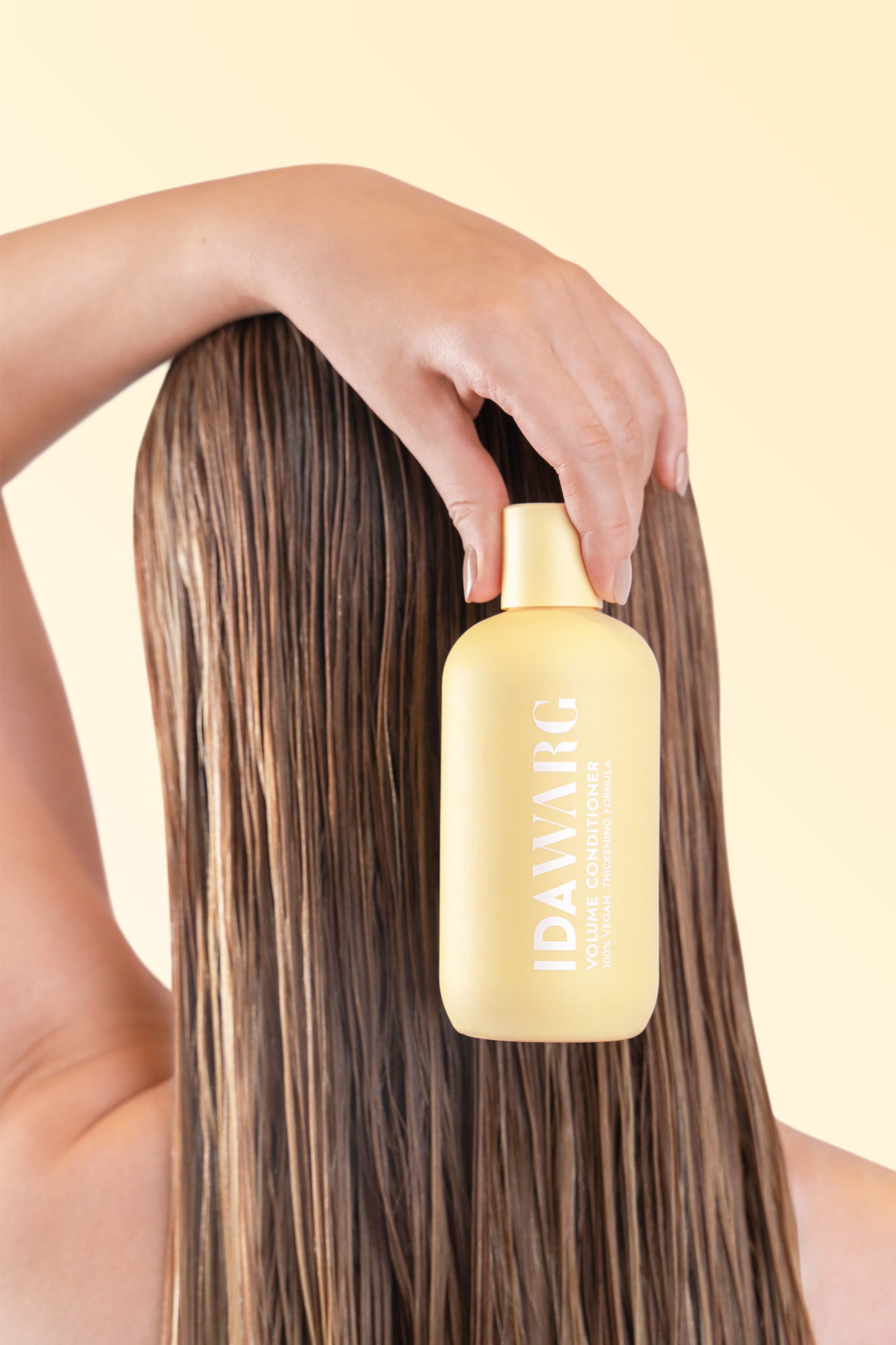 Größeres Bild ansehen: Die IDAWARG Volume Conditioner Flasche, ein hellgelber rechteckiger Behälter mit abgerundeten Kanten, wird von einer Hand aufrecht gehalten. Ihr vorderes Etikett, auf dem in weißer Schrift "IDAWARG VOLUME CONDITIONER 100% VEGAN. THICKENING FORMULA" steht, ist nach vorne gerichtet, neben langem, nassem Haar.