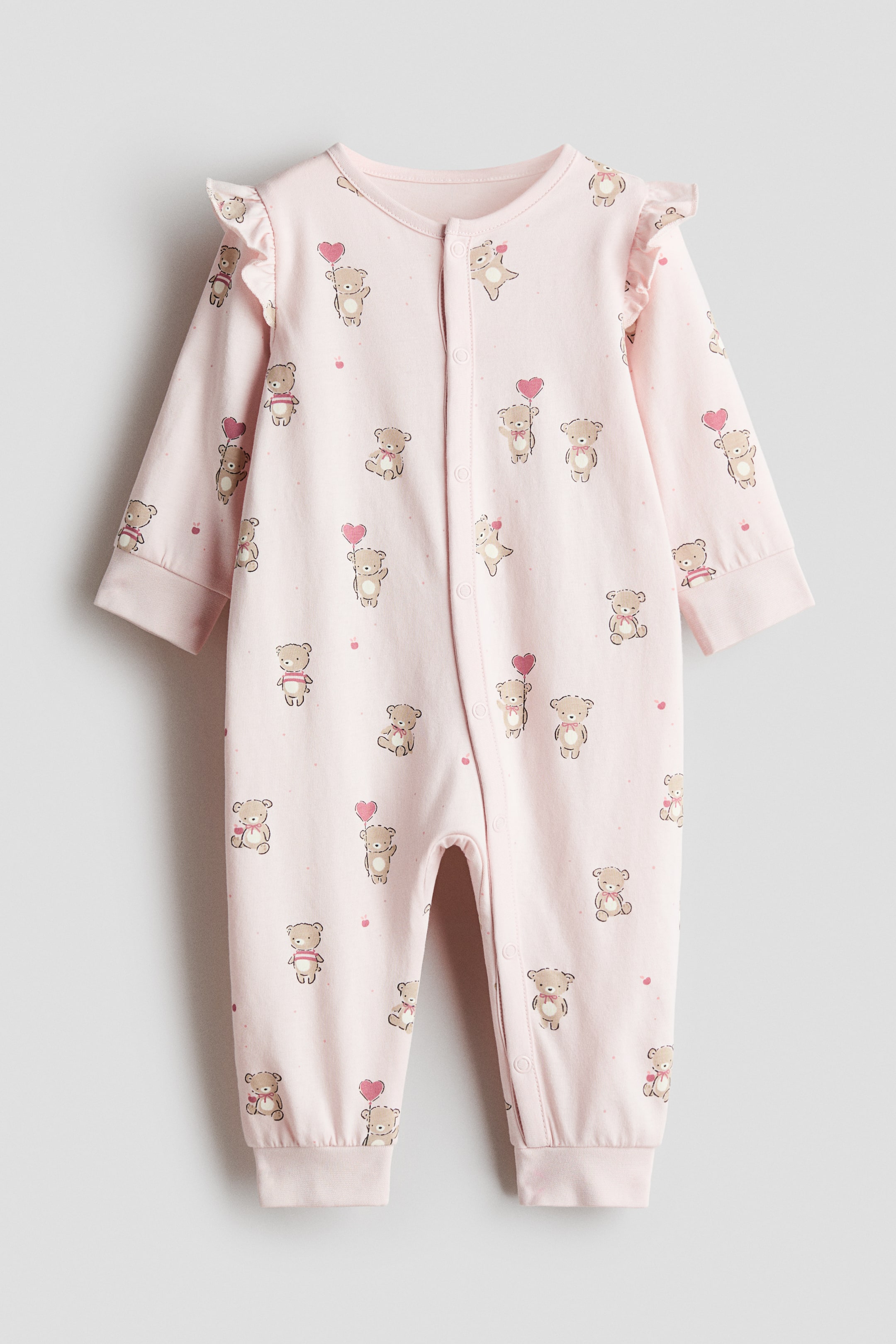 View larger image: A light pink sleepsuit in cotton jersey is lying flat, facing forward, and displaying a pattern of small teddy bears, some holding heart balloons. It is featuring ruffled frill trims over the shoulders, long sleeves with cuffs, long legs with cuffed hems, and press-studs running down the front and extending along one leg.