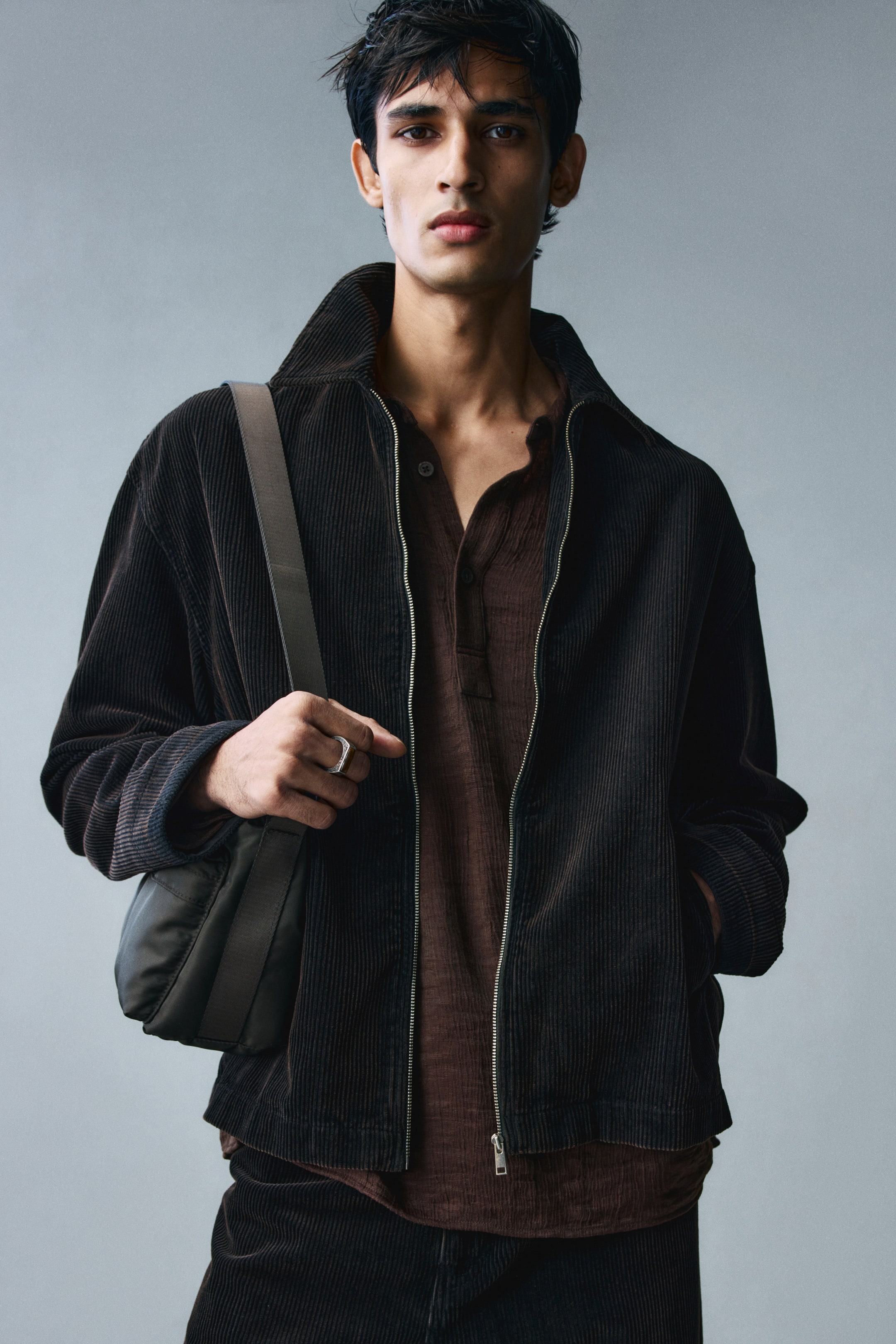 View larger image: A dark brown corduroy overshirt featuring a collar, front zipper, and straight hem is worn open by the model over a textured brown henley shirt. The model is holding the strap of a shoulder bag with one hand while resting the other hand inside the overshirt's front pocket.