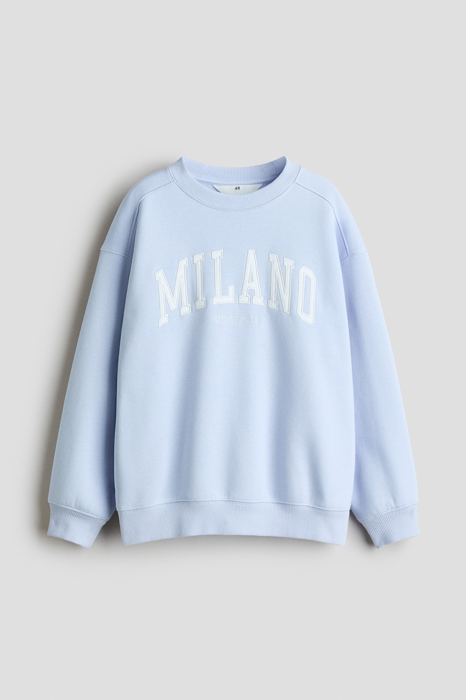 A light blue loose-fit sweatshirt is shown from the front, featuring a white printed 'MILANO SINCE 2011' design across the chest. The sweatshirt features a round, rib-trimmed neckline, dropped shoulders with forward-facing seams, and ribbed finishes at the cuffs and hem. - 1