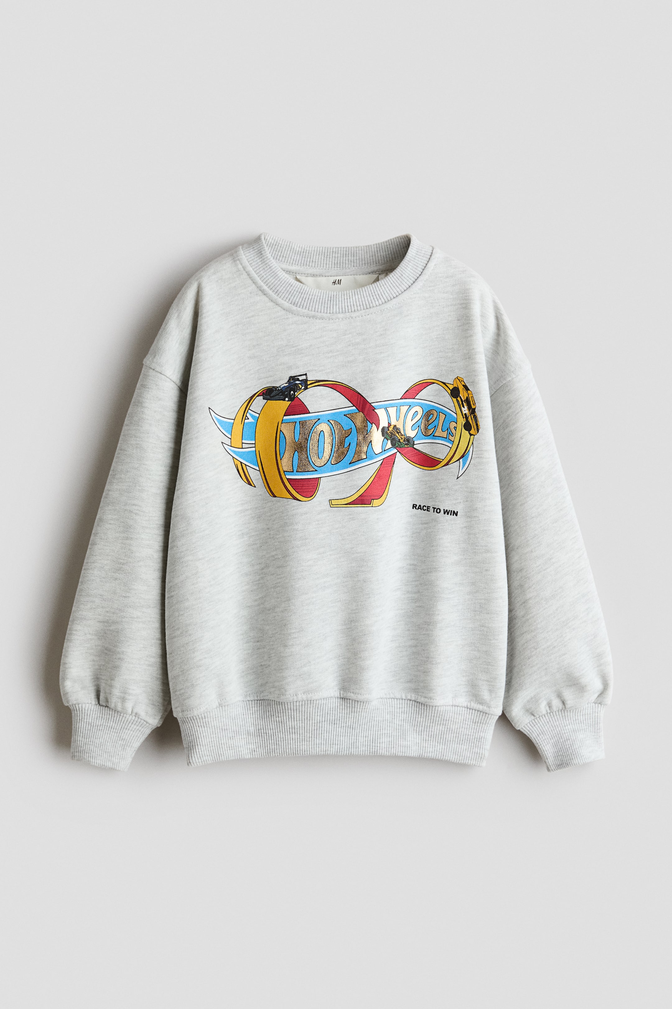 Agrandir l'image: Un sweat-shirt ample en maille chinée gris clair, présenté de face, avec un motif Hot Wheels coloré sur la poitrine et le texte « RACE TO WIN » imprimé en dessous. Le modèle est conçu avec un col rond bord-côte, des épaules tombantes et des manches longues terminées par des poignets côtelés, assortis à l’ourlet côtelé.