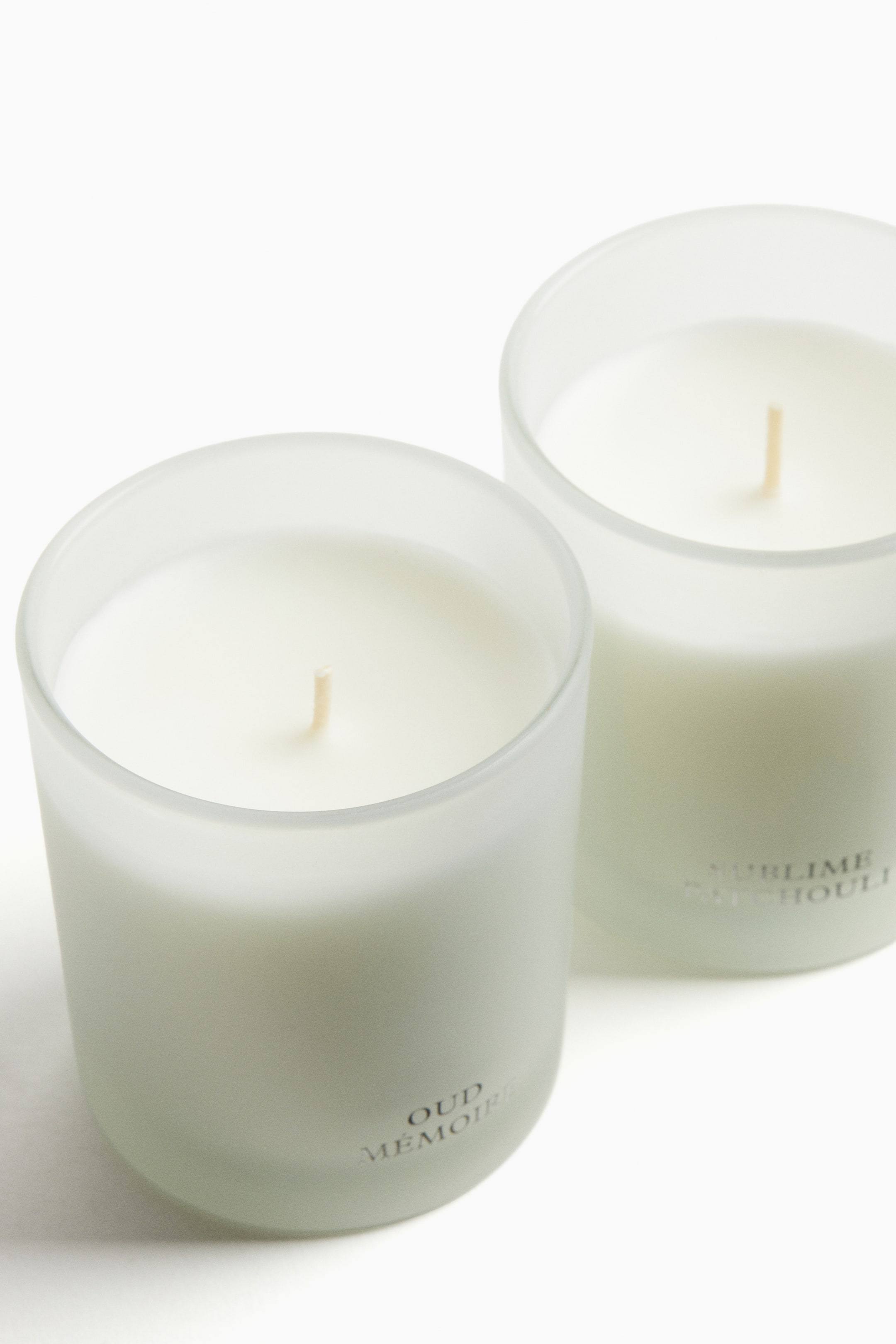 View larger image: Two candles are prominently displayed in frosted glass holders, shown from a slightly elevated front angle. Each holder is containing white wax with a centered wick, and the sides are featuring distinct text: "OUD MEMOIRE" on the front candle and "SUBLIME PATCHOULI" on the back.