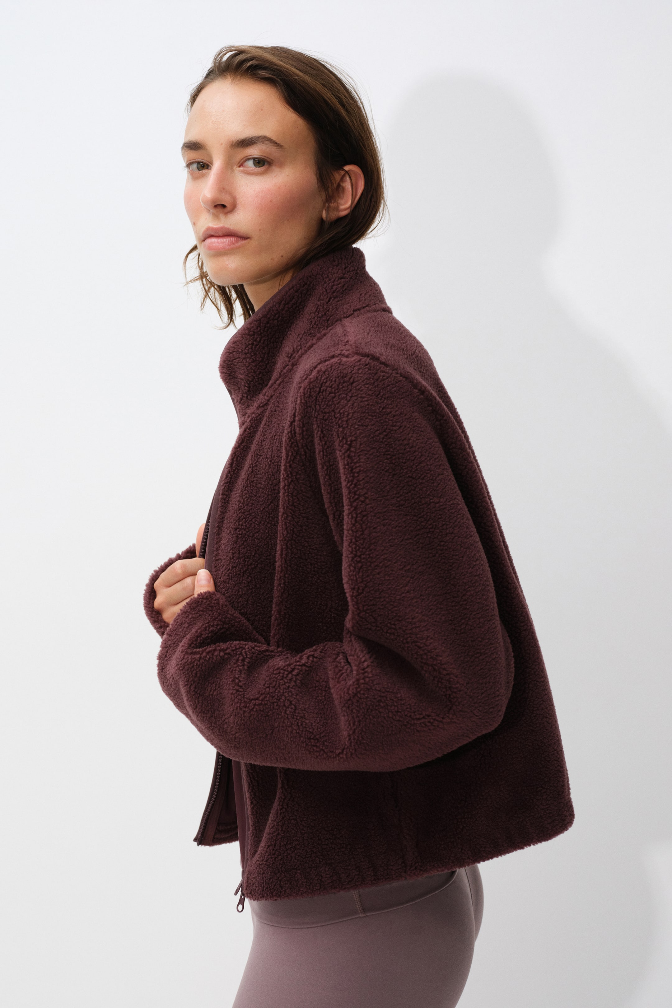 View larger image: A dark burgundy fleece jacket with a high collar and front zipper is worn by the model, who is holding the front opening with one hand. The jacket is styled with matching mauve leggings visible at the waist.