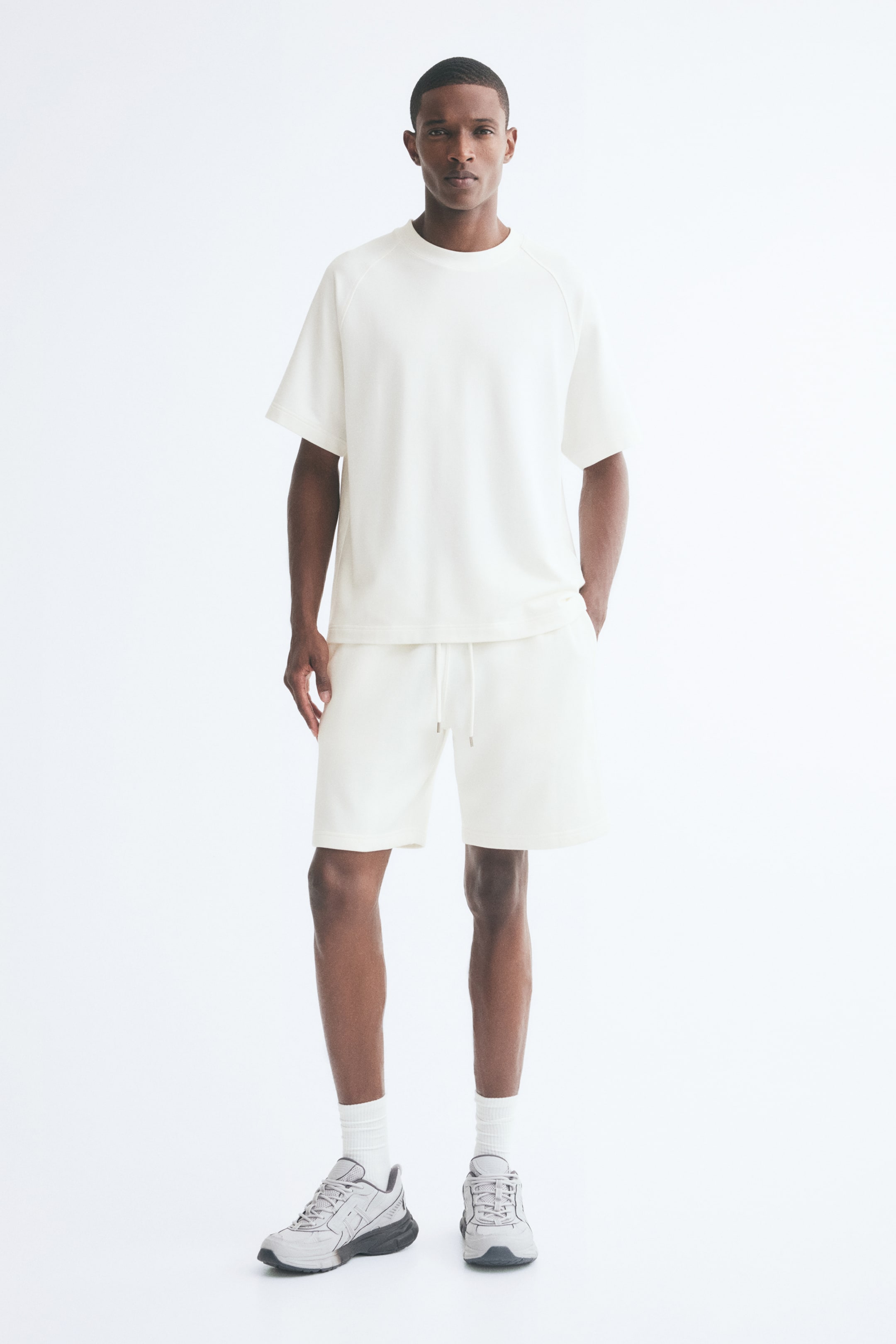 View larger image: Off-white sweat shorts in a regular fit with an elasticated waist and hanging drawstrings are worn by the model, who is standing with one hand resting in a side pocket. The shorts are styled with a matching short-sleeved T-shirt, white crew socks, and grey sneakers.