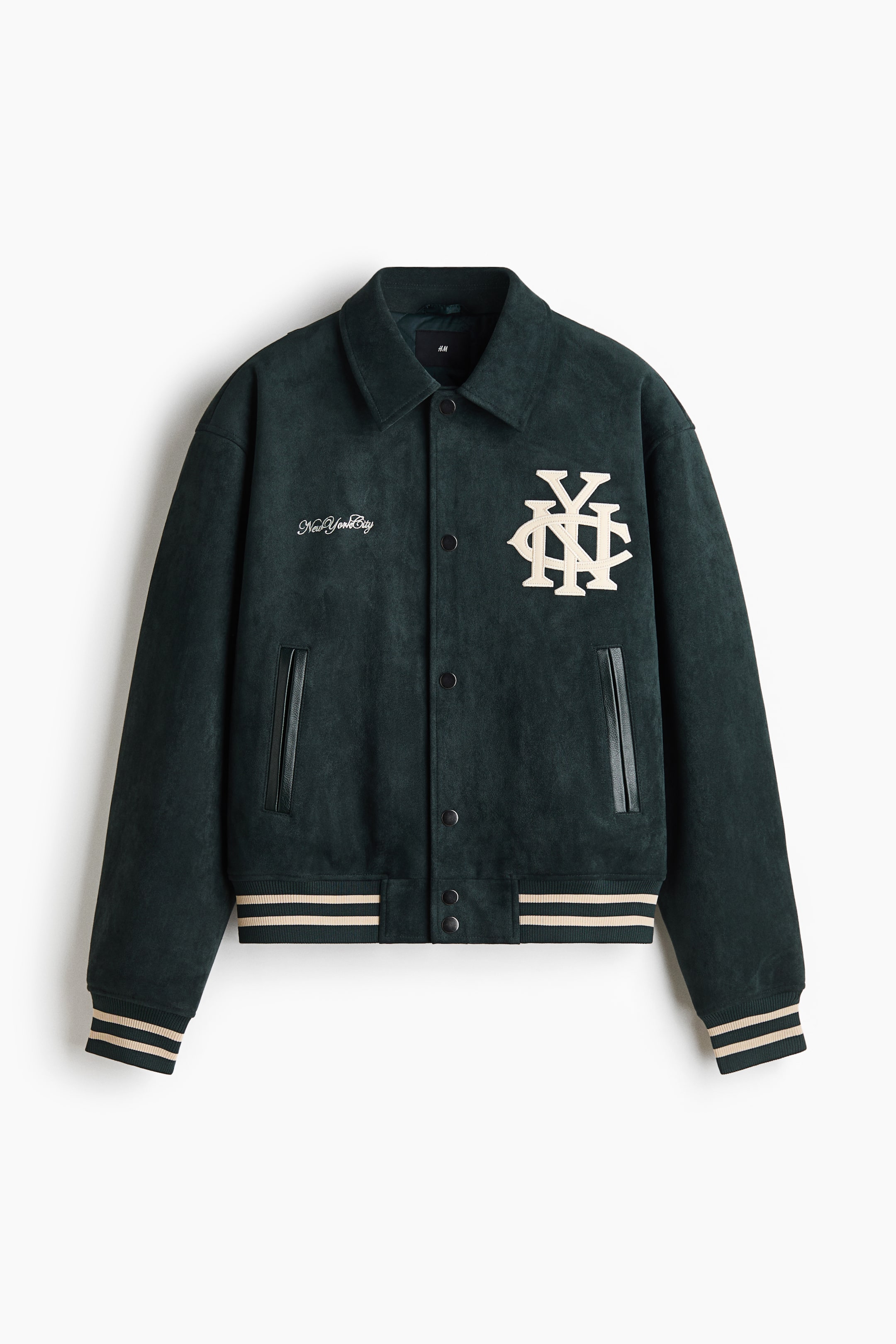 View larger image: A dark green, suede-textured varsity jacket is shown from the front, featuring a classic spread collar, a snap-button front closure, and ribbed cuffs and hem accented with cream-coloured stripes. The jacket is detailed with a large cream monogram patch on the left chest, a small script embroidery on the right chest, and vertical welt pockets with black trim.