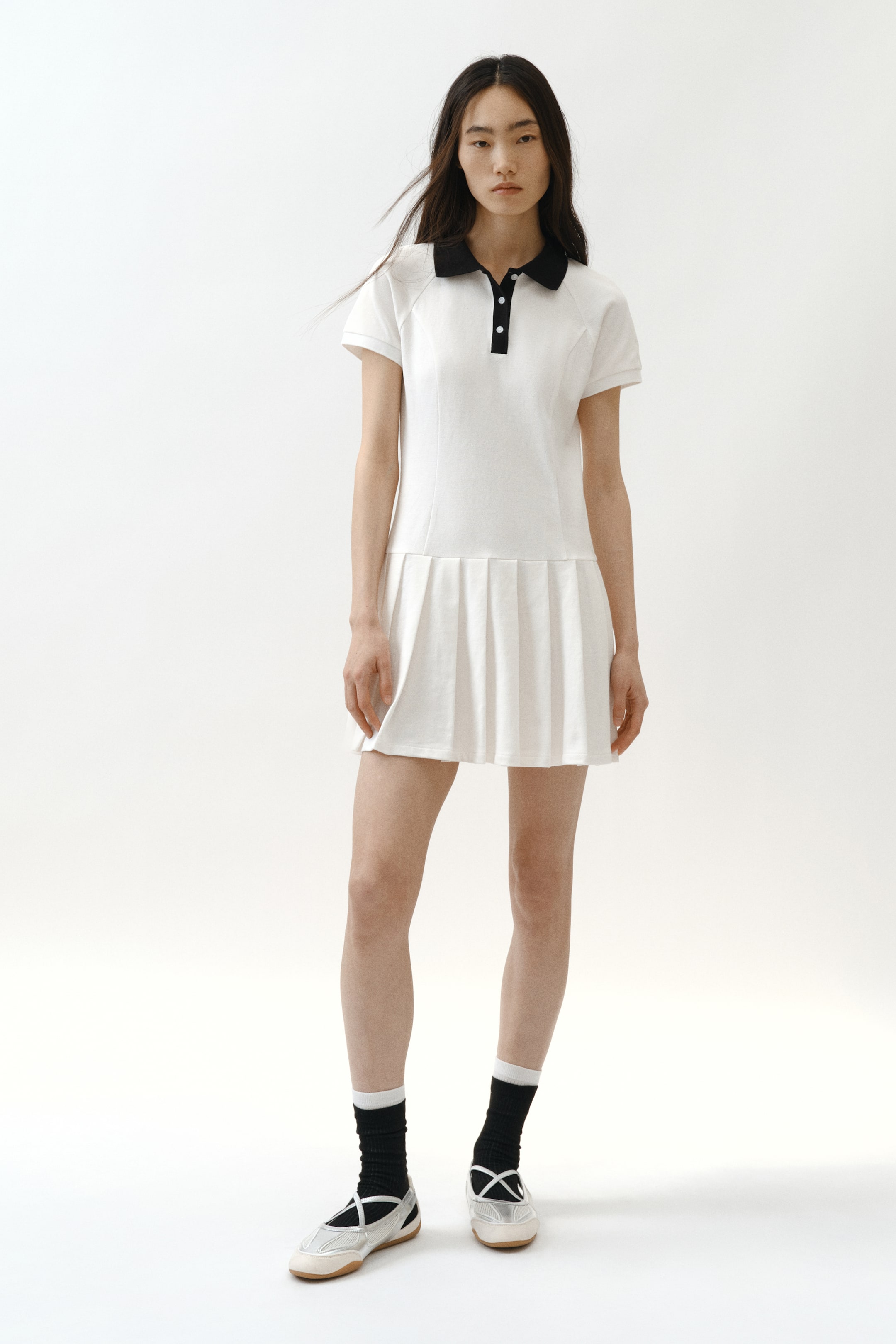 拡大画像を表示: 白いテニススタイルのポロドレスは、黒いポロカラー、黒い三つボタンの前立て、ラグランショートスリーブ、プリーツ入りのミニスカートが特徴です。