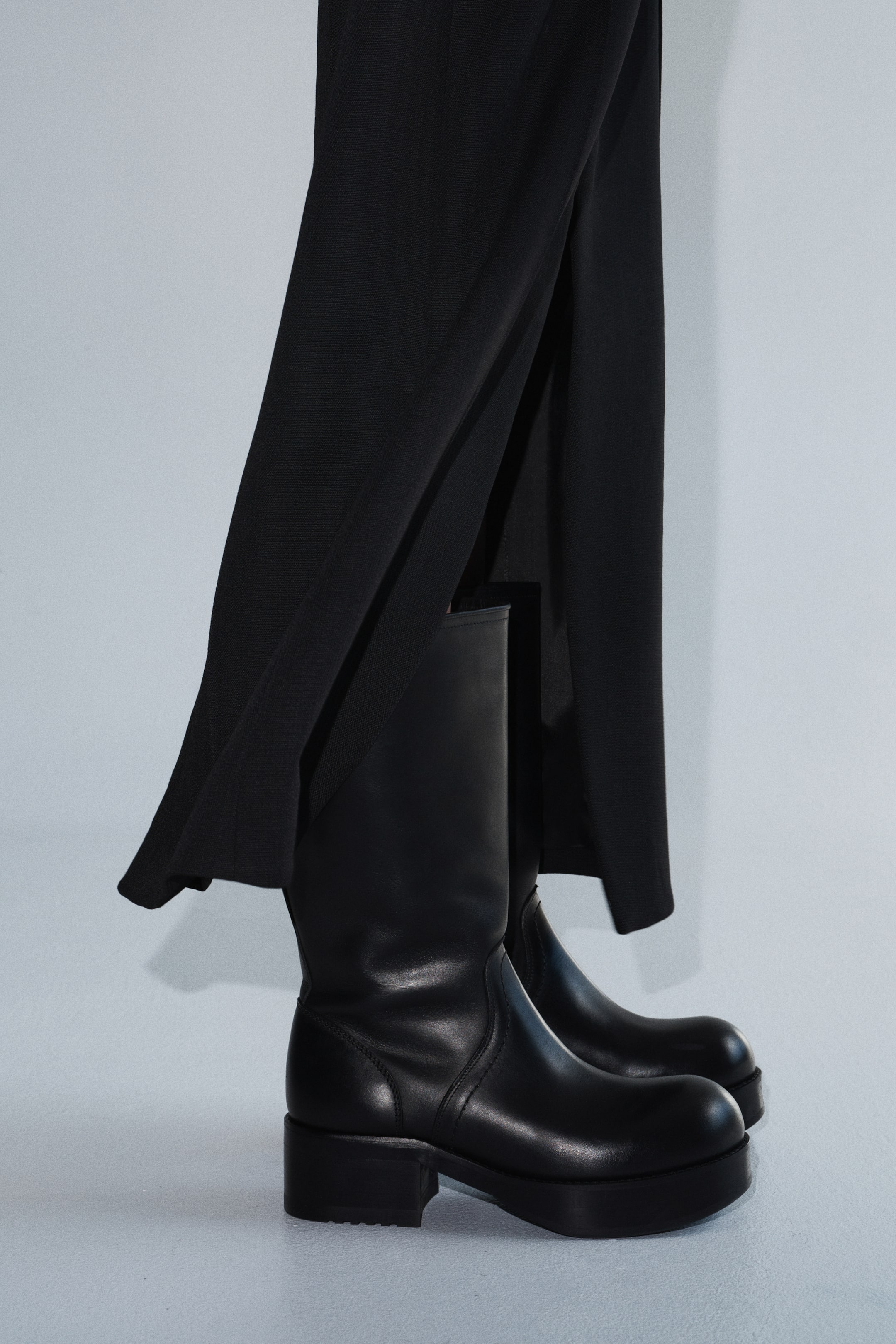 Visualizza l'immagine ingrandita: La modella indossa stivali neri in pelle al polpaccio. Presentano una spessa suola platform e un tacco a blocco, con pantaloni neri che coprono parzialmente i gambali degli stivali.