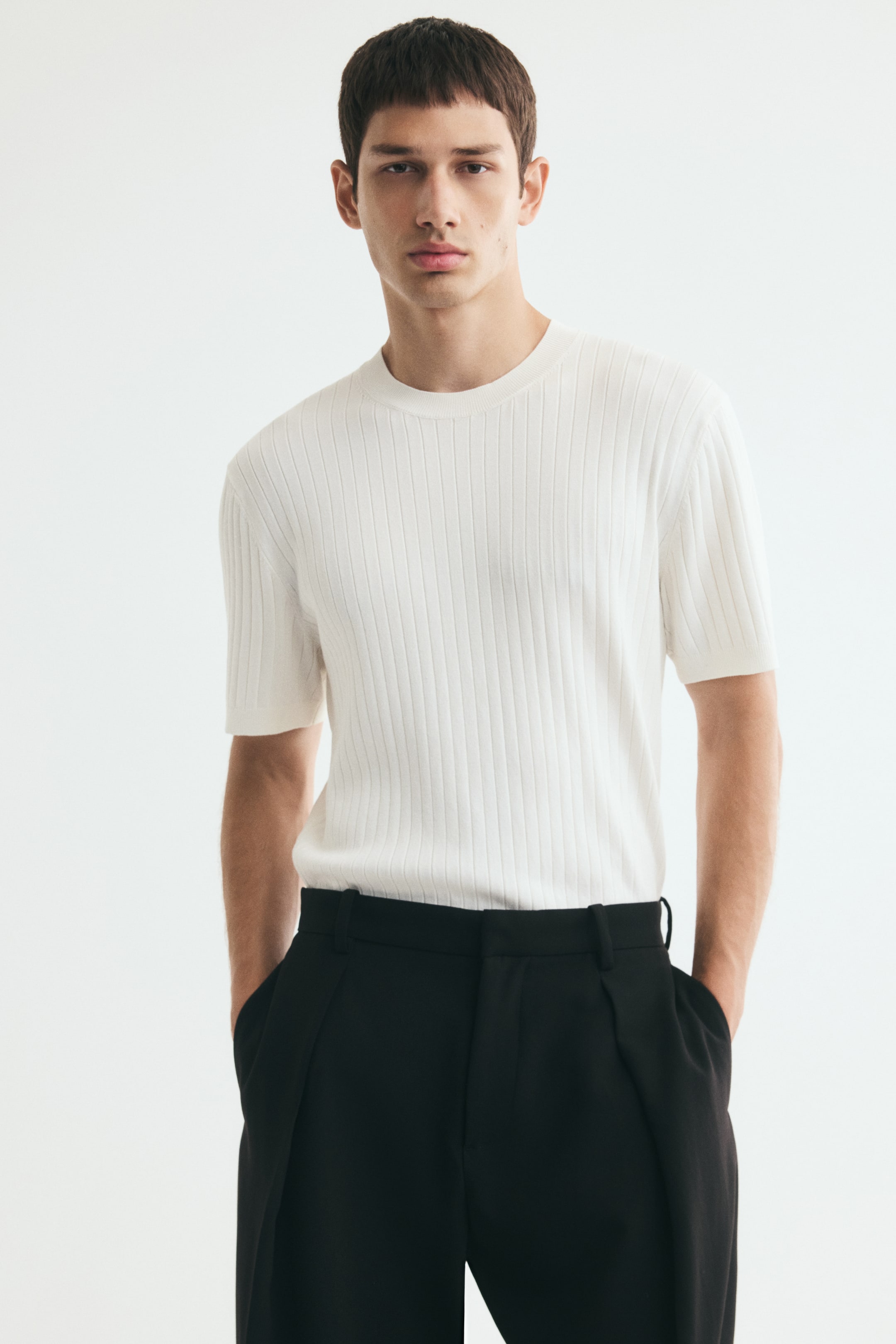 View larger image: The model is wearing a cream-coloured fine rib-knit tee with a round neckline and short sleeves. The slim-fit shirt is tucked into black trousers, accentuating its fitted silhouette.