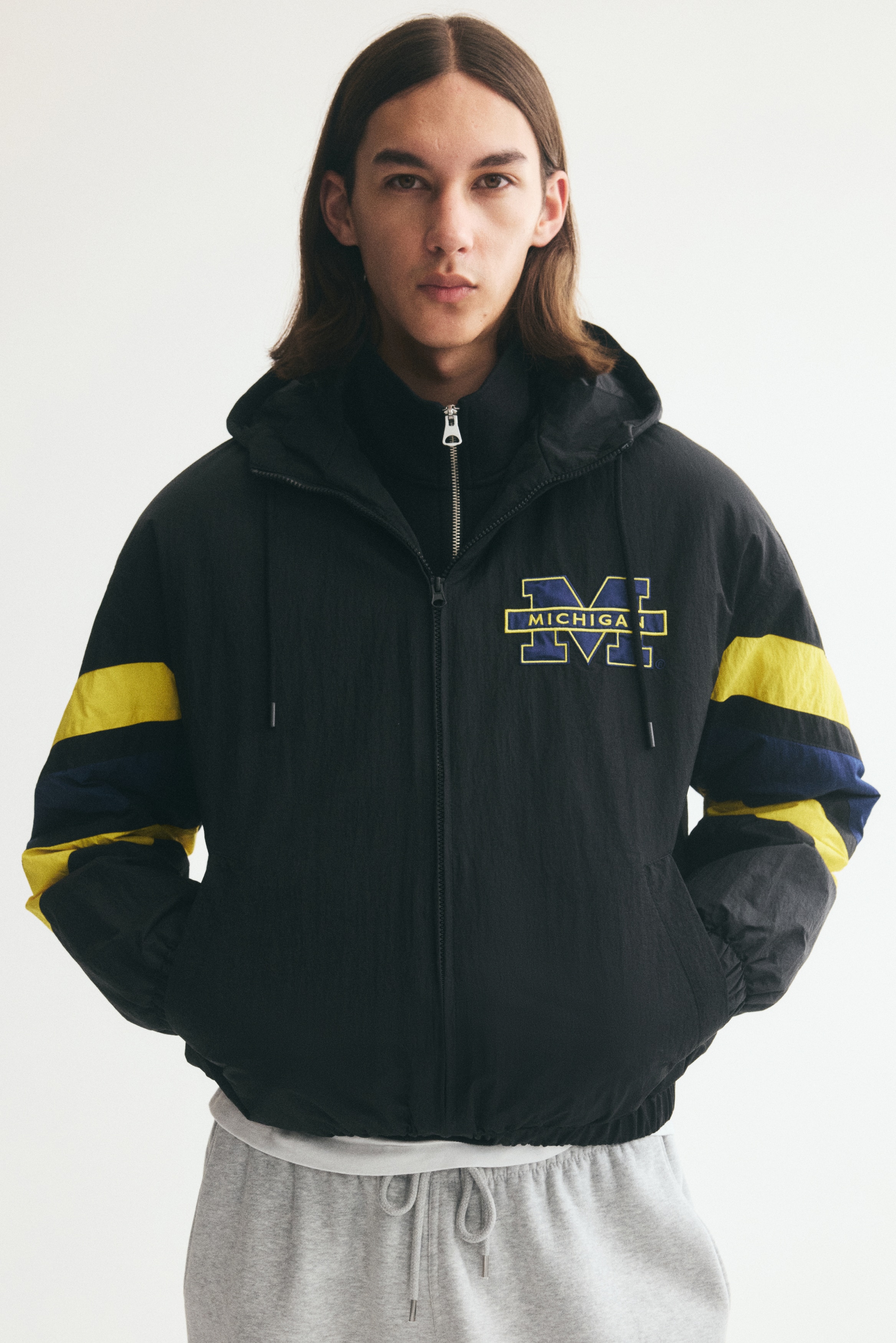 This black hooded windbreaker jacket is shown from the front, featuring a drawstring hood, a full-length zip fastening, slanted welt pockets, and an elasticated hem and cuffs. It displays a blue and yellow "MICHIGAN" logo on the left chest, with thick diagonal yellow and blue stripes wrapping around the sleeves.