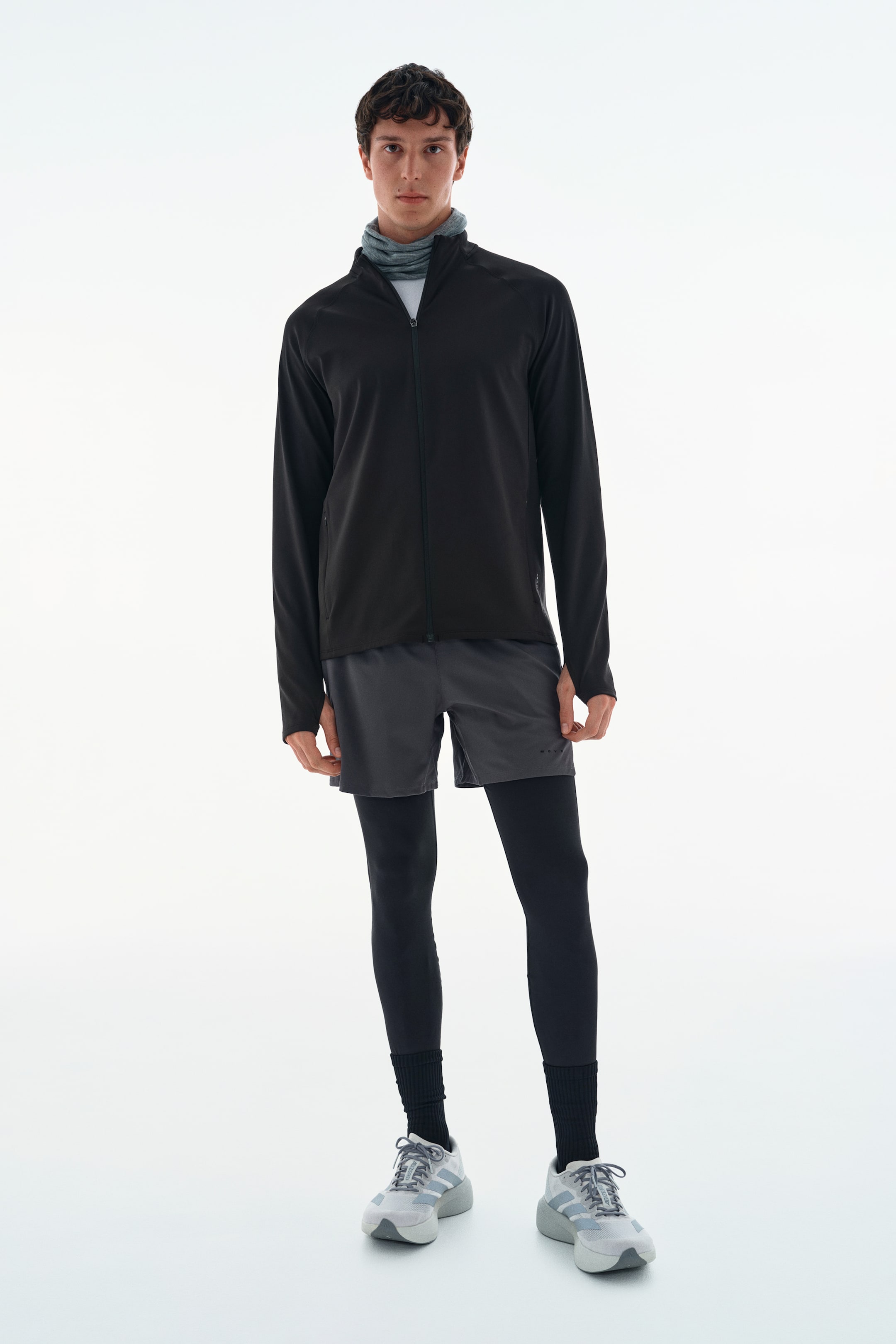 View larger image: A black, long-sleeved athletic jacket with a zippered front and thumbholes is being worn by the model. The jacket is styled with a grey neck gaiter, dark grey shorts layered over black leggings, and grey athletic shoes.