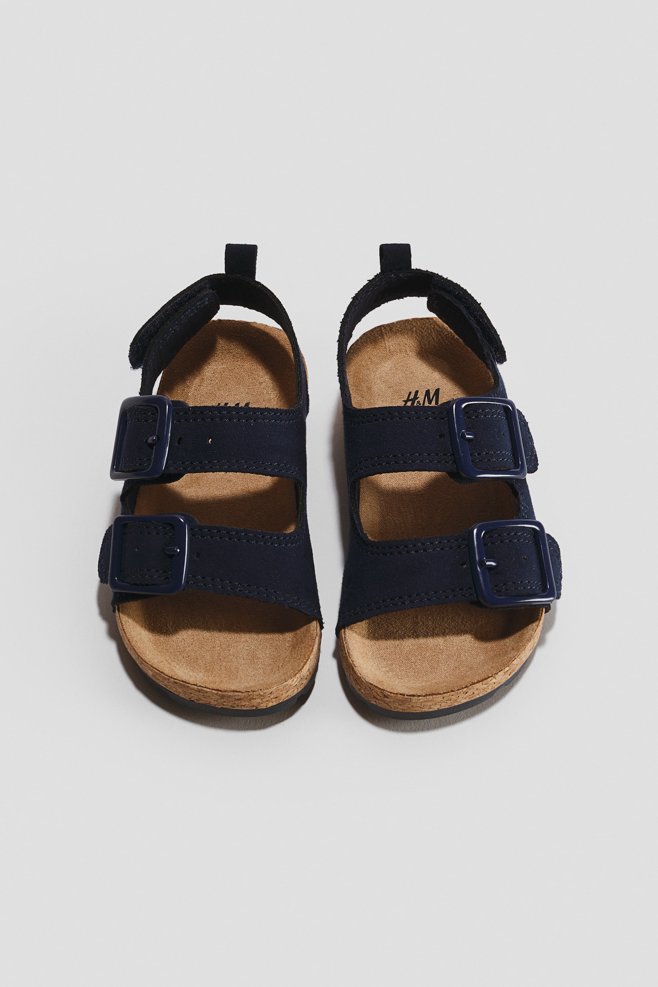 View larger image: A pair of navy blue sandals is shown from a front, top-down angle, featuring two adjustable foot straps with matching blue buckles and an ankle strap with a hook-and-loop fastening. The sandals are designed with moulded tan insoles, a pull-loop at the back, and dark outsoles.