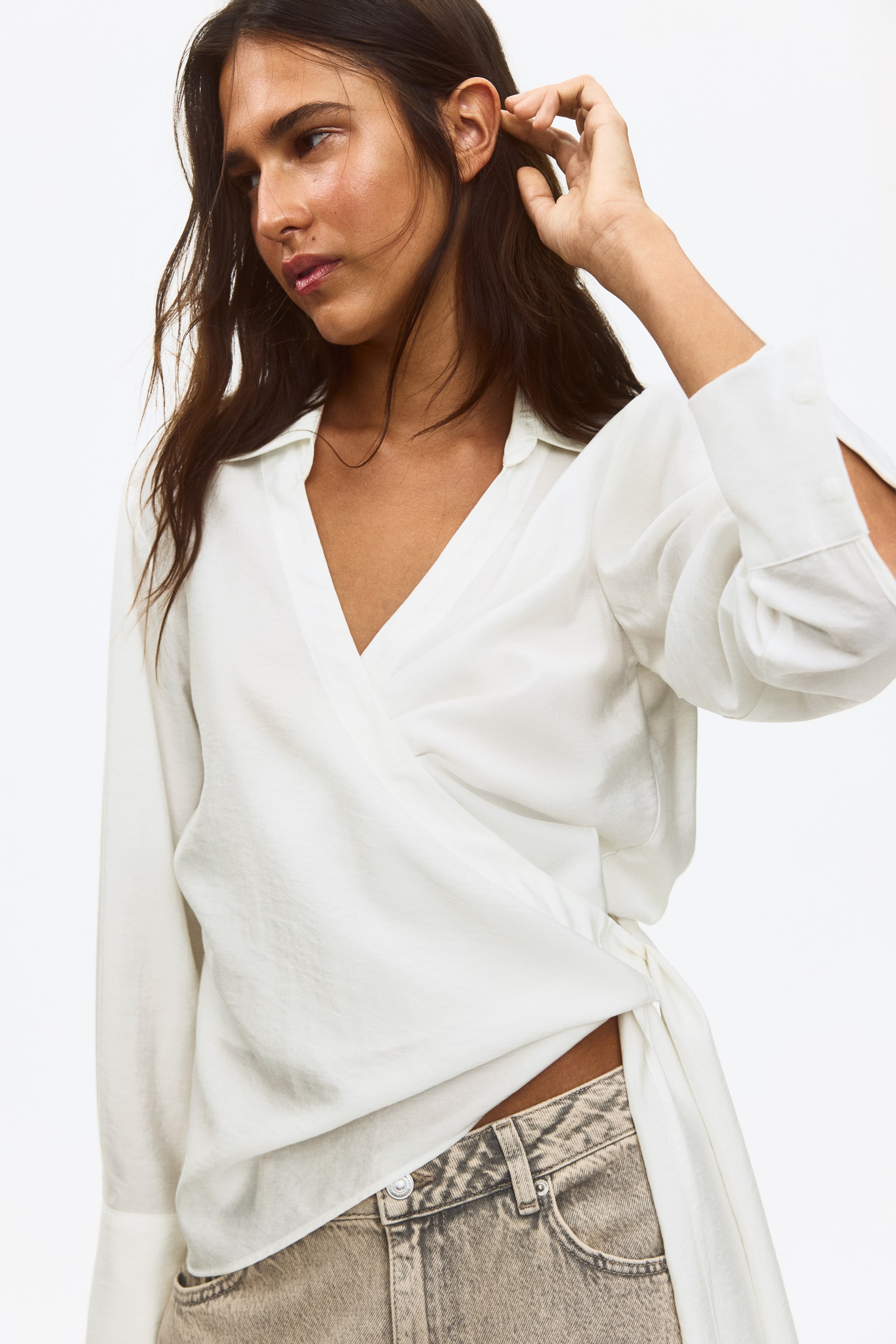 View larger image: The model is wearing a white viscose-blend wrapover shirt, showcasing an open collar, subtly dropped shoulders, and long sleeves with buttoned cuffs. The wrapover front is secured by wide ties around the waist, tied at the side, with an asymmetric hem draping over light-colored jeans.
